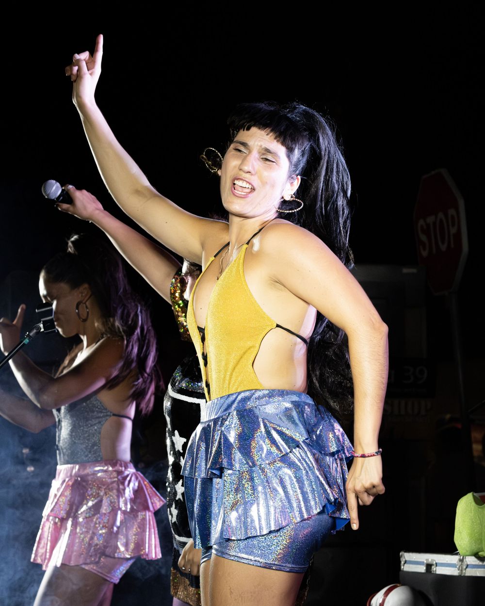 A woman in a yellow halter top and silver skirt performs energetically on stage, raising her hand with a stop sign behind her.