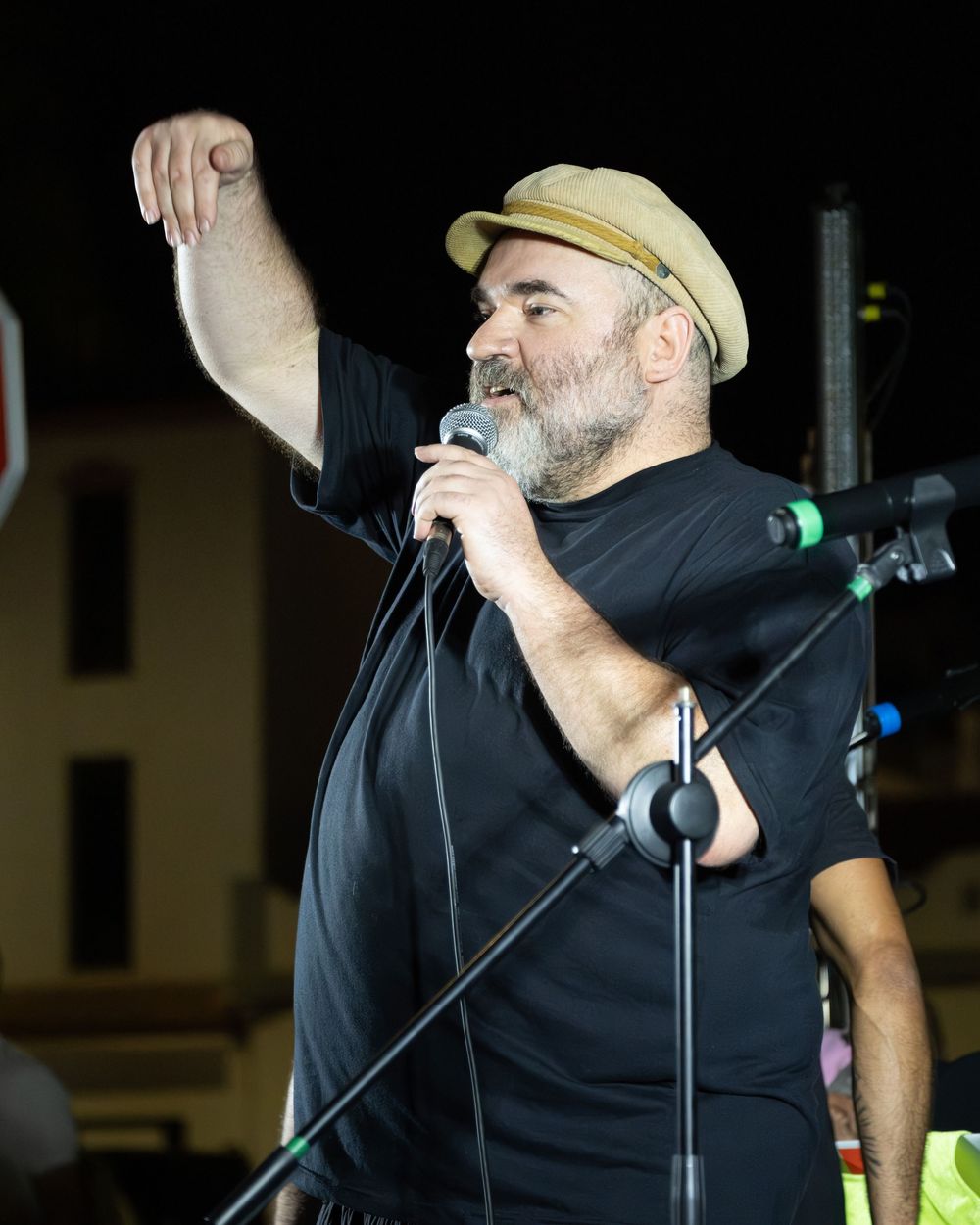 Middle-aged man with gray beard and tan flat cap speaking into microphone while gesturing at outdoor event.