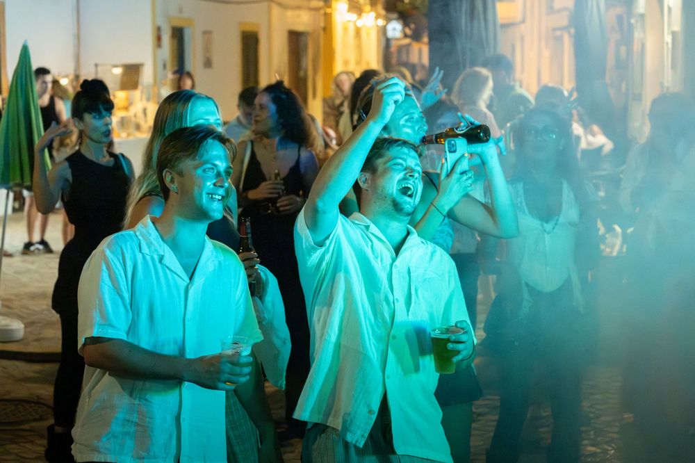 Two friends in white shirts laugh and take photos at a vibrant nighttime outdoor party with teal lighting.