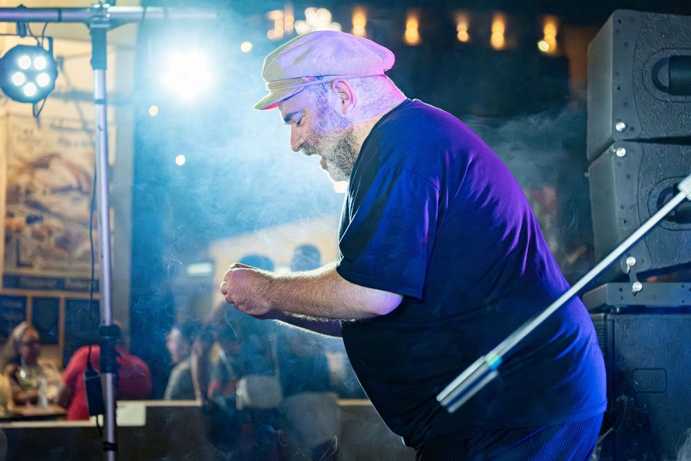 Bearded man in blue t-shirt and cap performing on stage under dramatic blue and purple stage lighting.