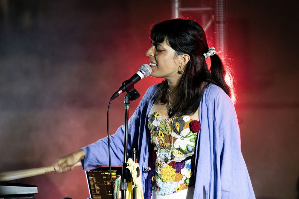A woman singer performs on stage with a microphone, wearing a floral dress and blue kimono under colorful stage lights.