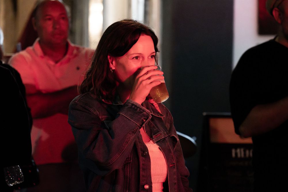 Woman in denim jacket drinking beer at a dimly lit bar with red ambient lighting.