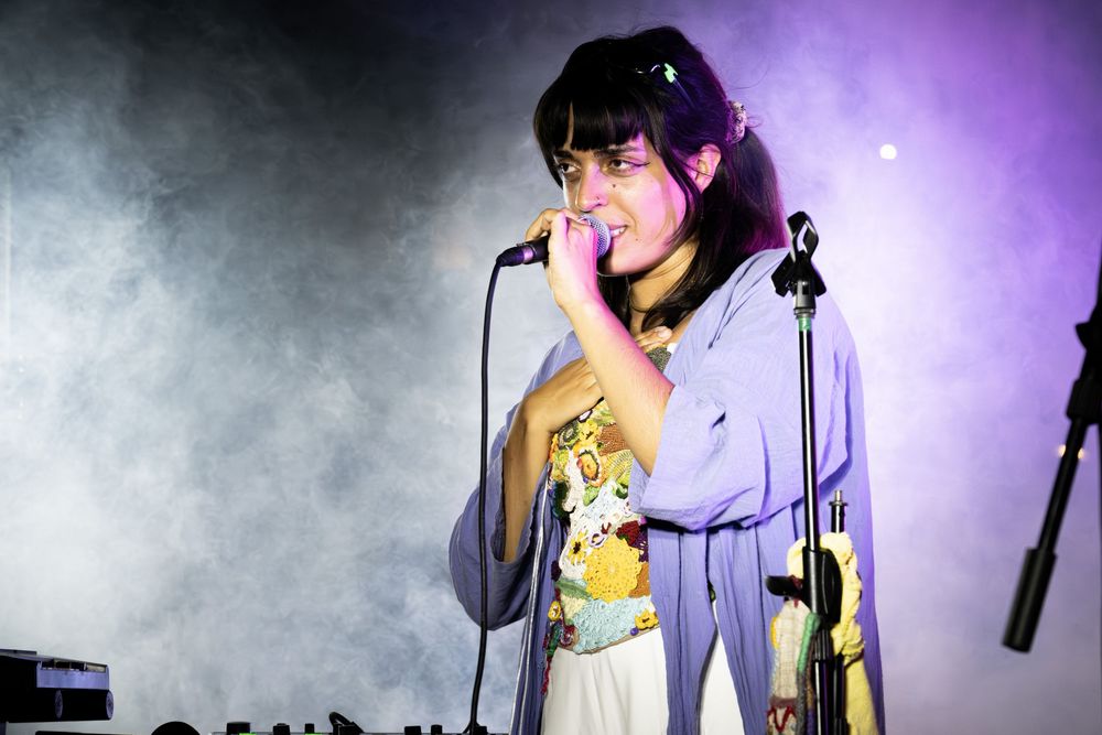 Young woman singer performing on stage with microphone, wearing lavender outfit under dramatic stage lighting.