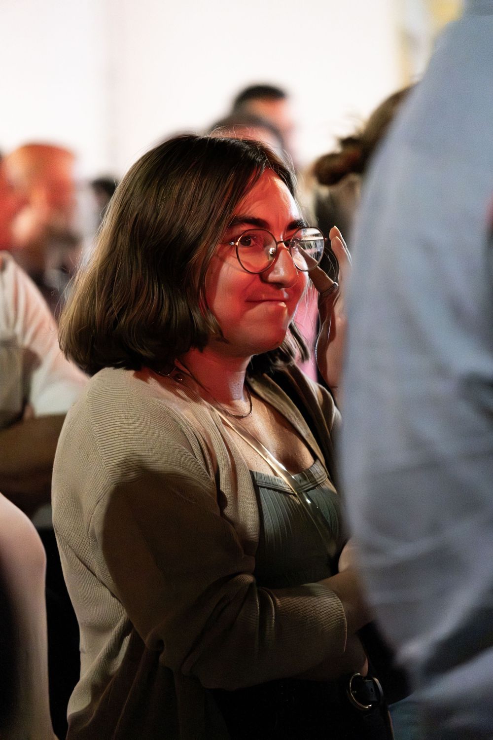 Young woman with round glasses and brown hair stands with arms crossed at an indoor event.