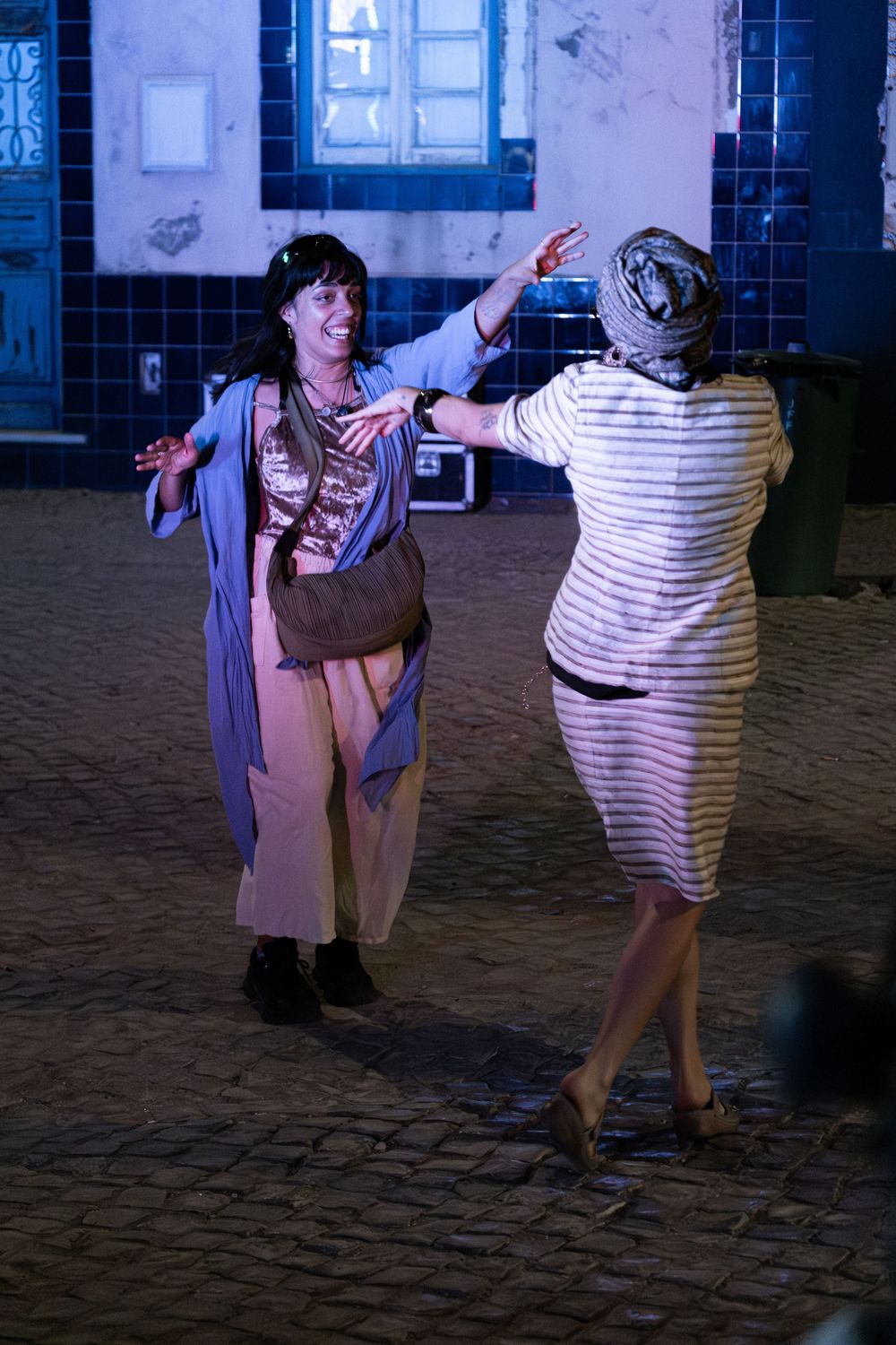 Two women dancing together on a cobblestone street at night near a tiled building facade.