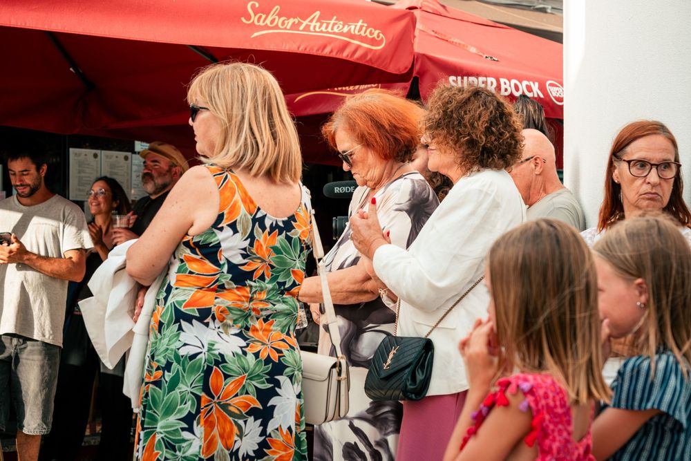 Crowd of people gathered outdoors at a summer event near a red Super Bock umbrella on a sunny day.