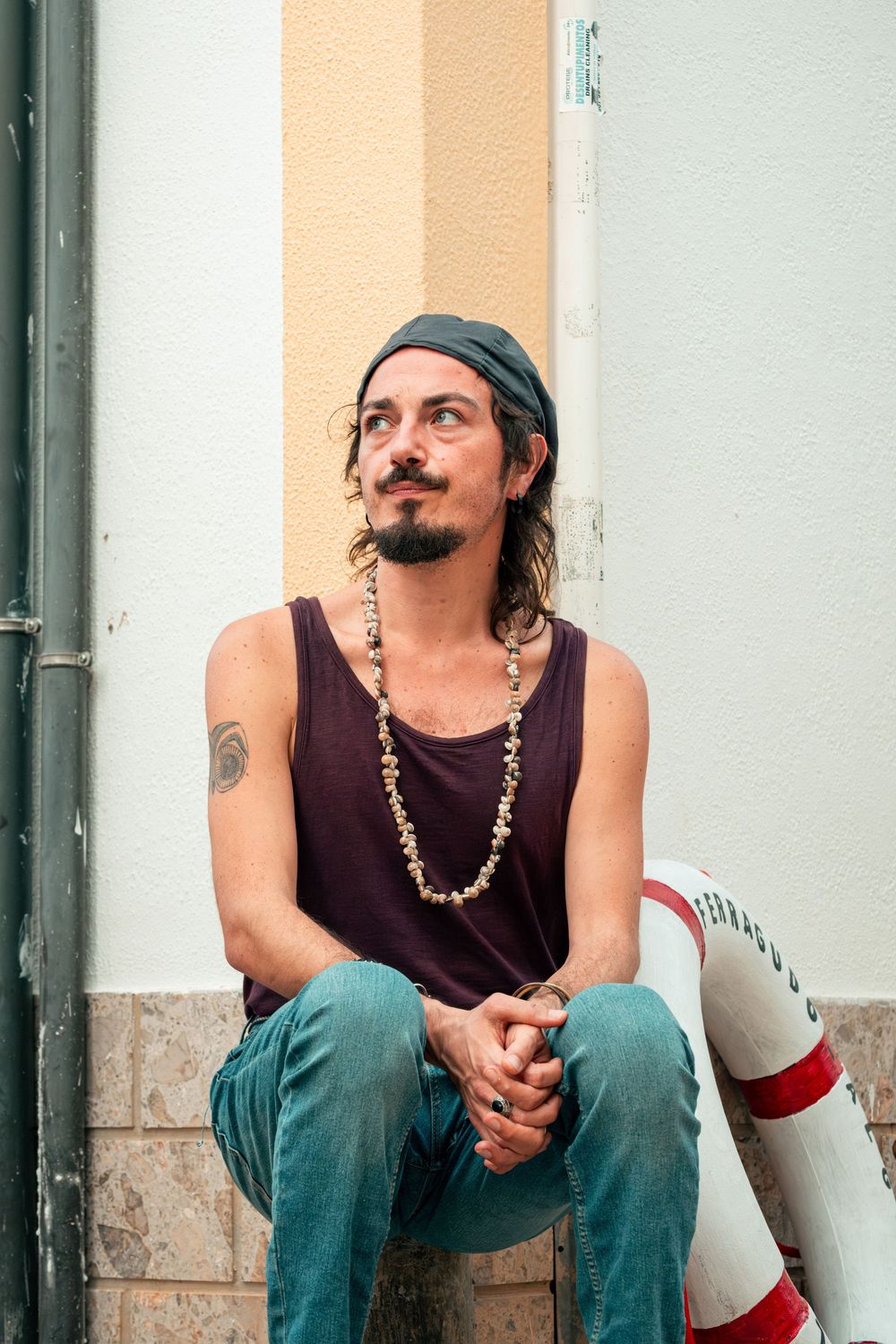 A bohemian man with a bandana, bead necklace, and tattoos sits outdoors in a tank top and jeans, gazing thoughtfully upward.
