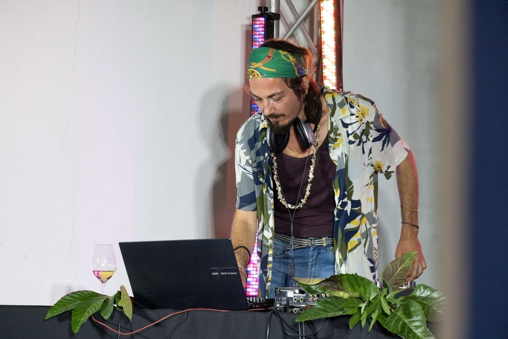 DJ in tropical shirt and green headband mixing music on a laptop at an outdoor event.