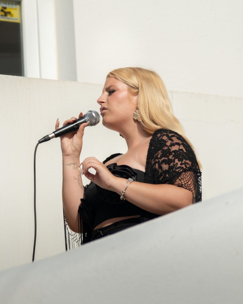 Blonde woman in black lace top singing into microphone at outdoor event.
