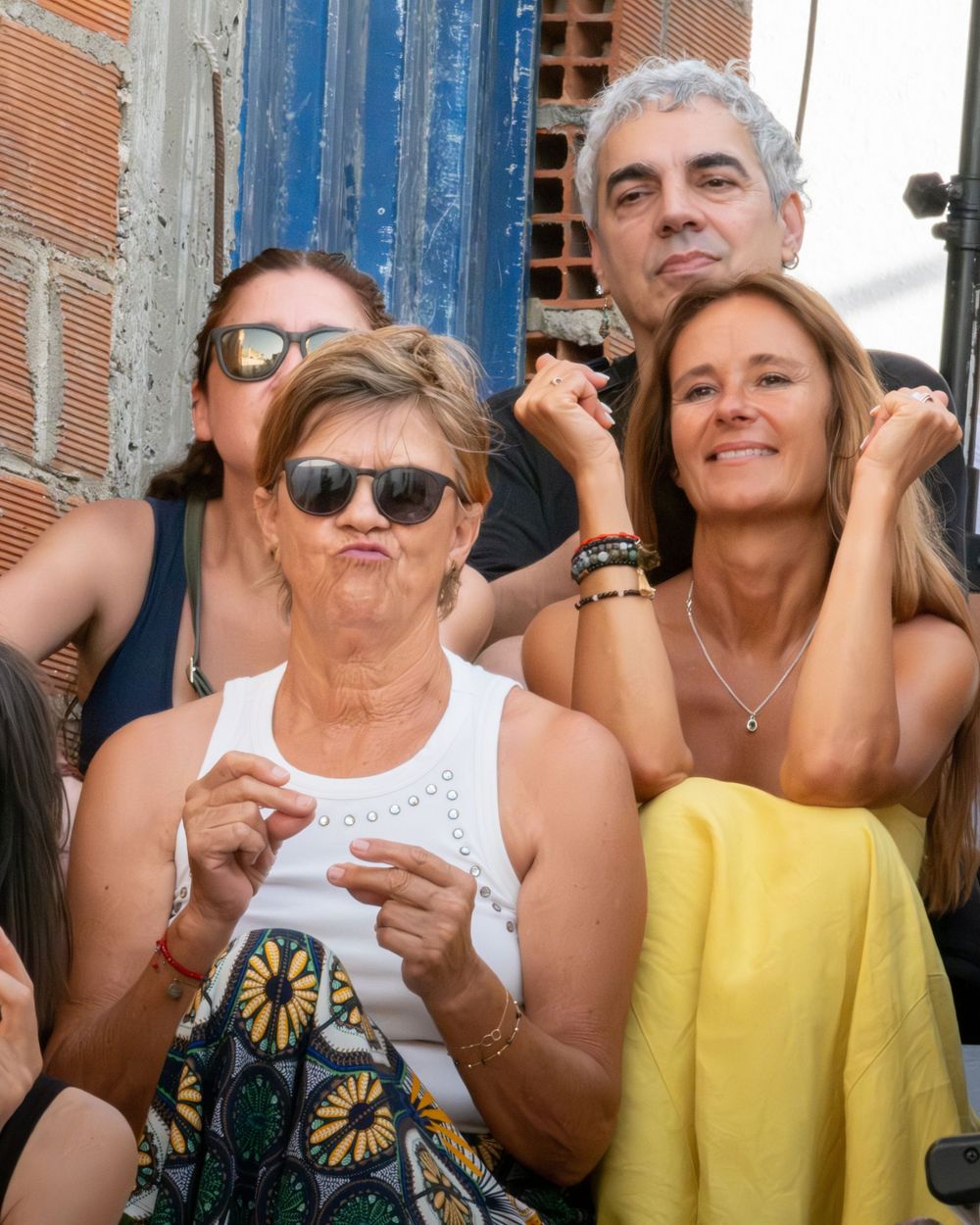 Group of people enjoying an outdoor event, woman in white tank top making funny face, another woman in yellow skirt smiling.