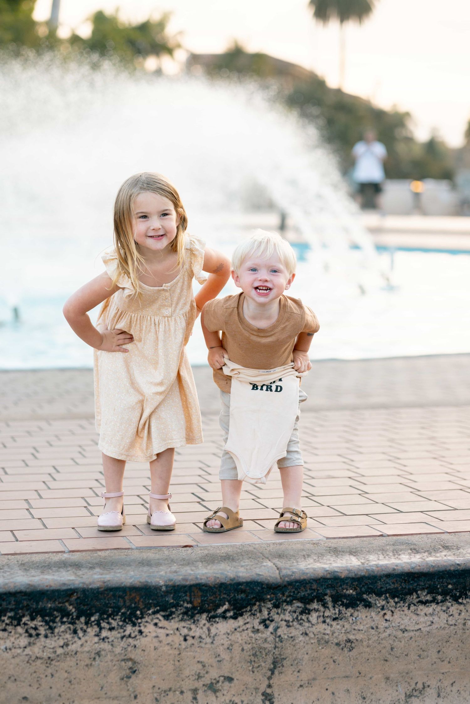 Two young siblings smiling together near a fountain in warm evening light.