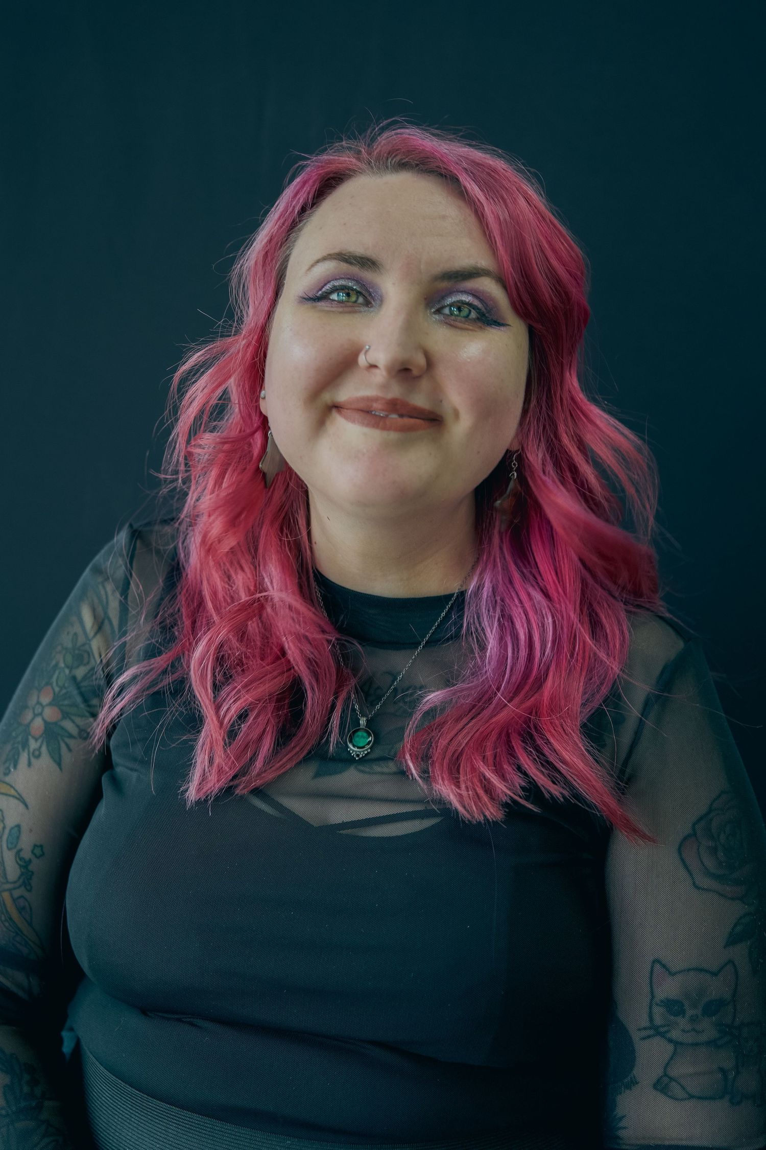 Woman with pink hair, tattoos, and green eyes smiling, wearing black mesh top and teal necklace.