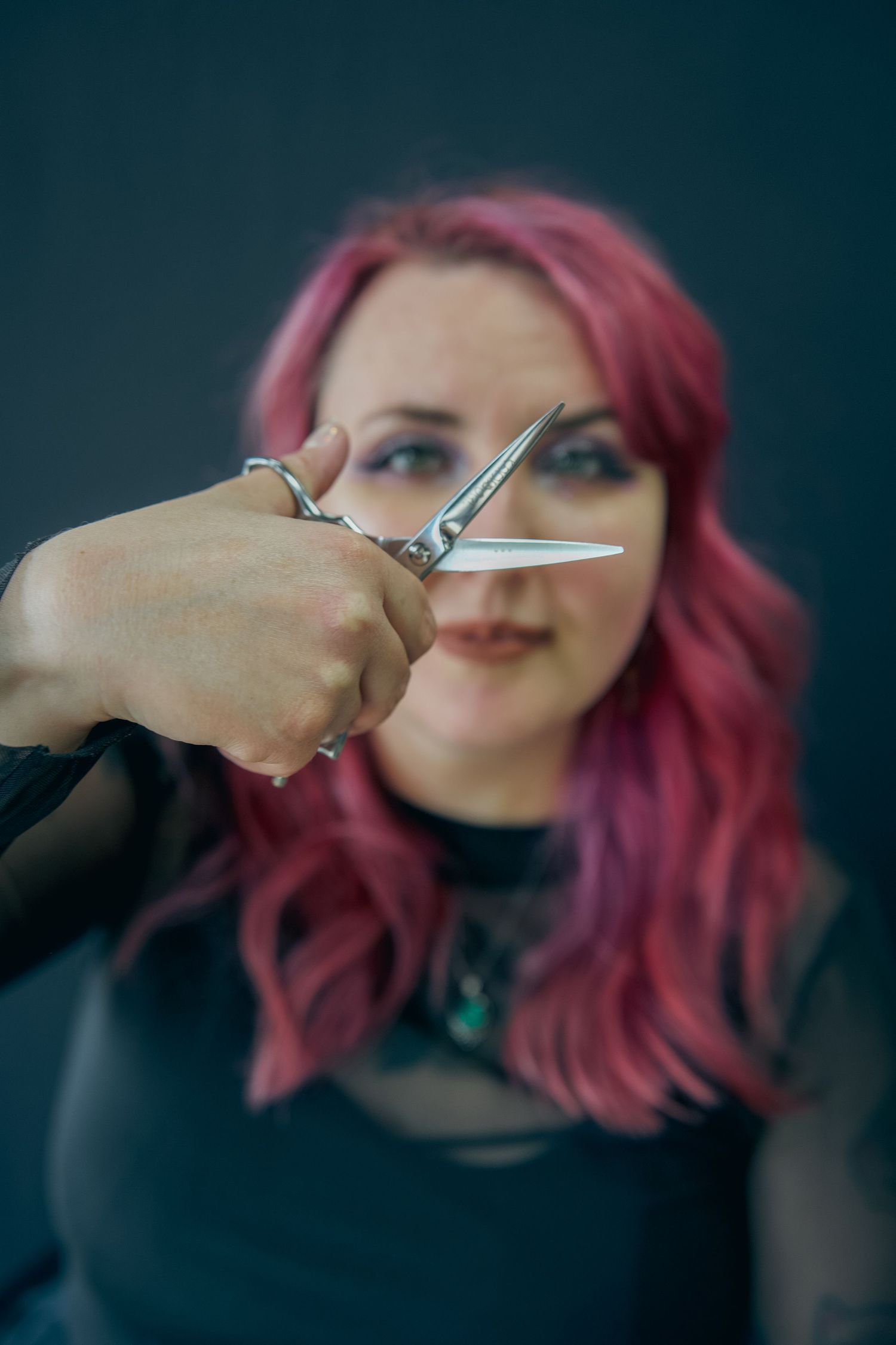 A woman with pink hair holds scissors toward the camera in a hair salon setting.