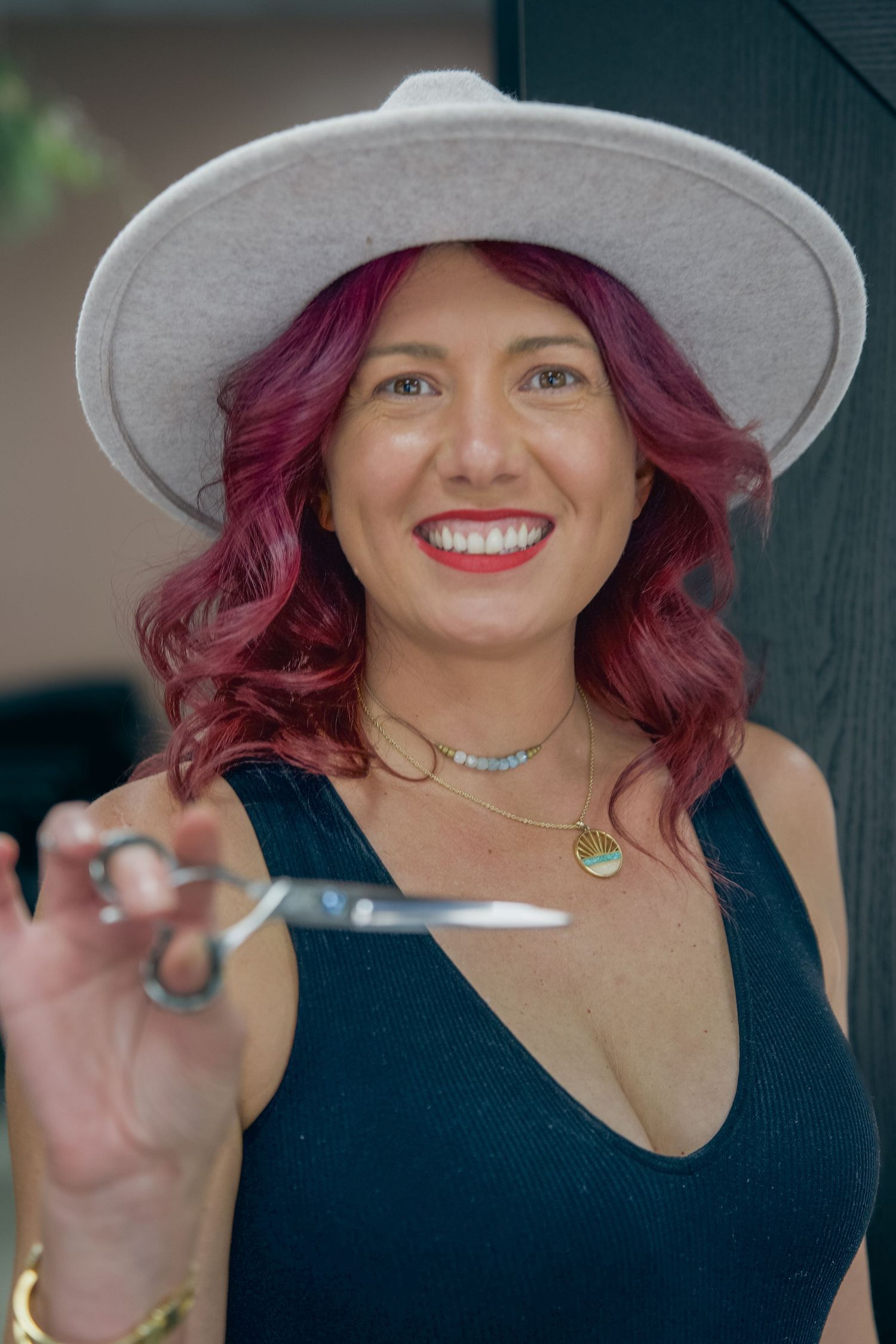 Smiling woman with red hair and white hat holds scissors, wearing a teal top and gold necklace, likely a hairstylist.