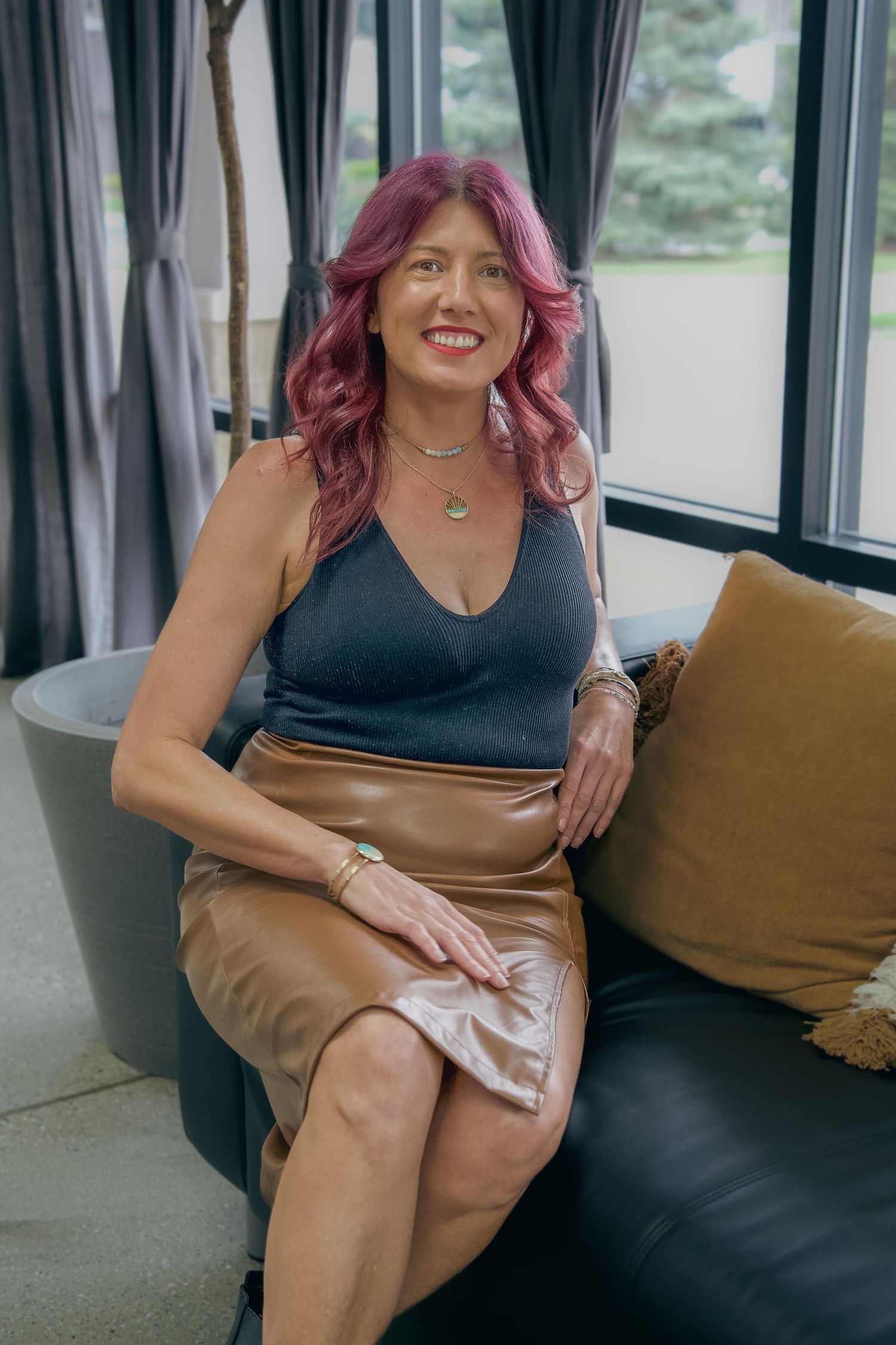 Woman with pink hair in black top and brown leather skirt sitting on dark sofa smiling indoors.