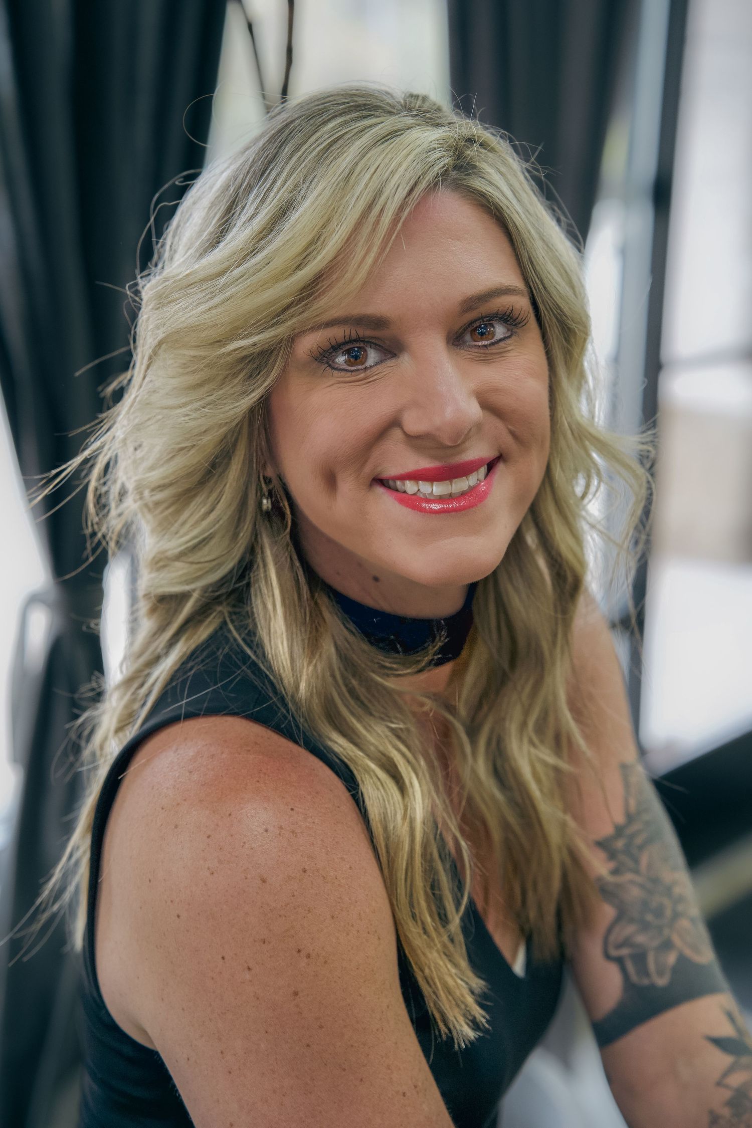 Smiling blonde woman with red lips, choker necklace, and arm tattoo wearing black sleeveless top.