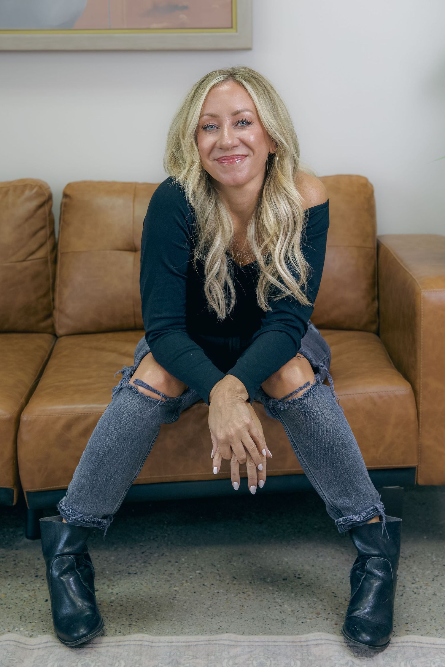 Blonde woman in black top and ripped jeans sitting on brown leather couch with black boots.