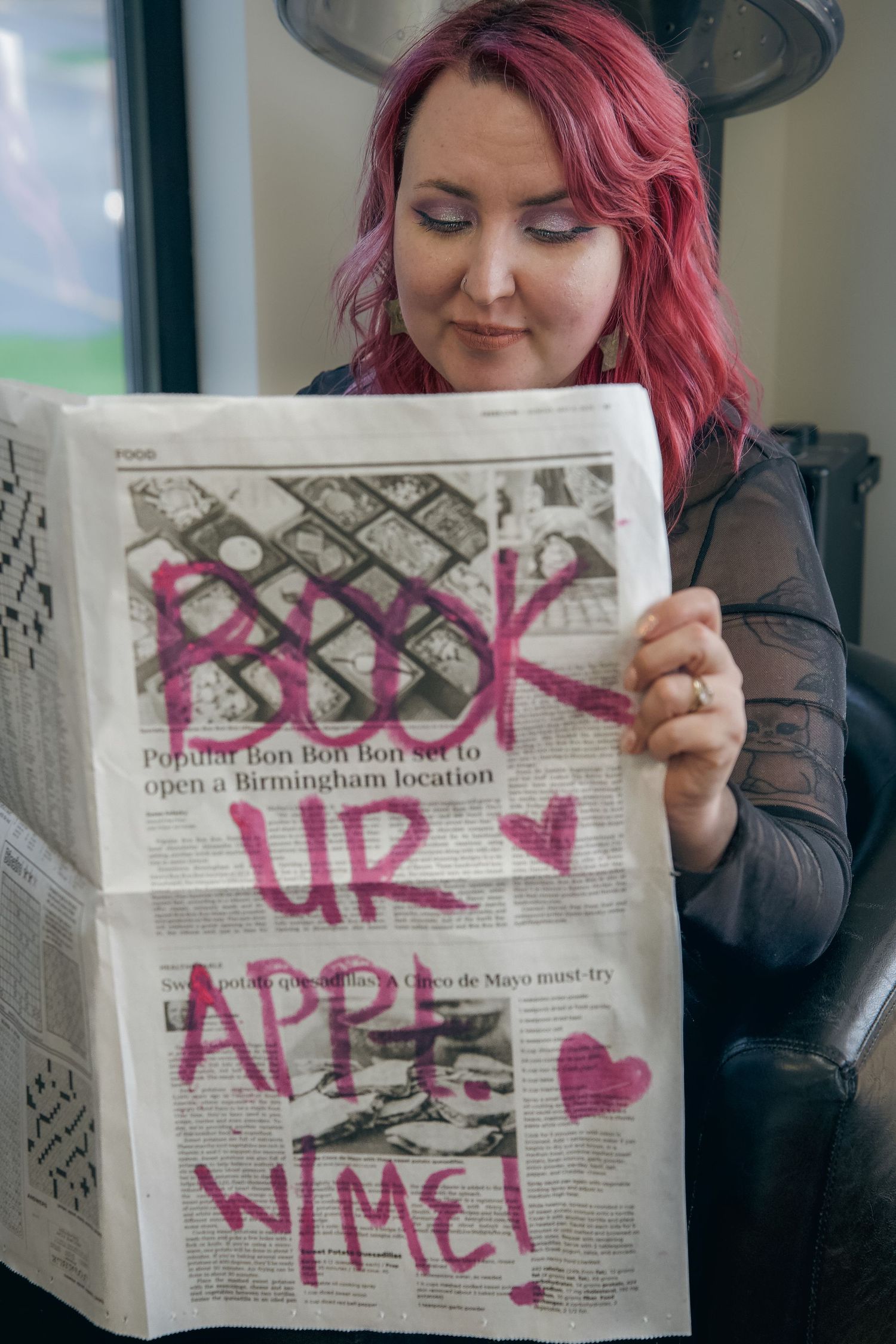 Woman with pink hair reading a newspaper with pink graffiti text saying Book UR Appt W Me at a salon.
