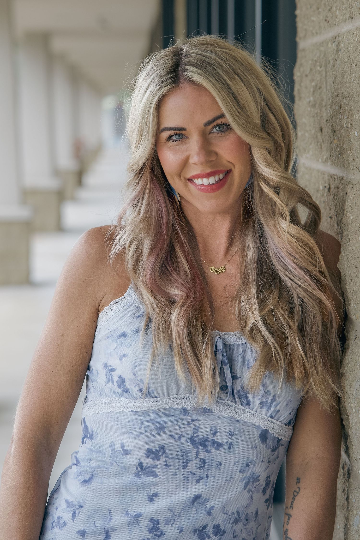 Smiling blonde woman in blue floral sundress with pink highlights leaning against a stone column outdoors.