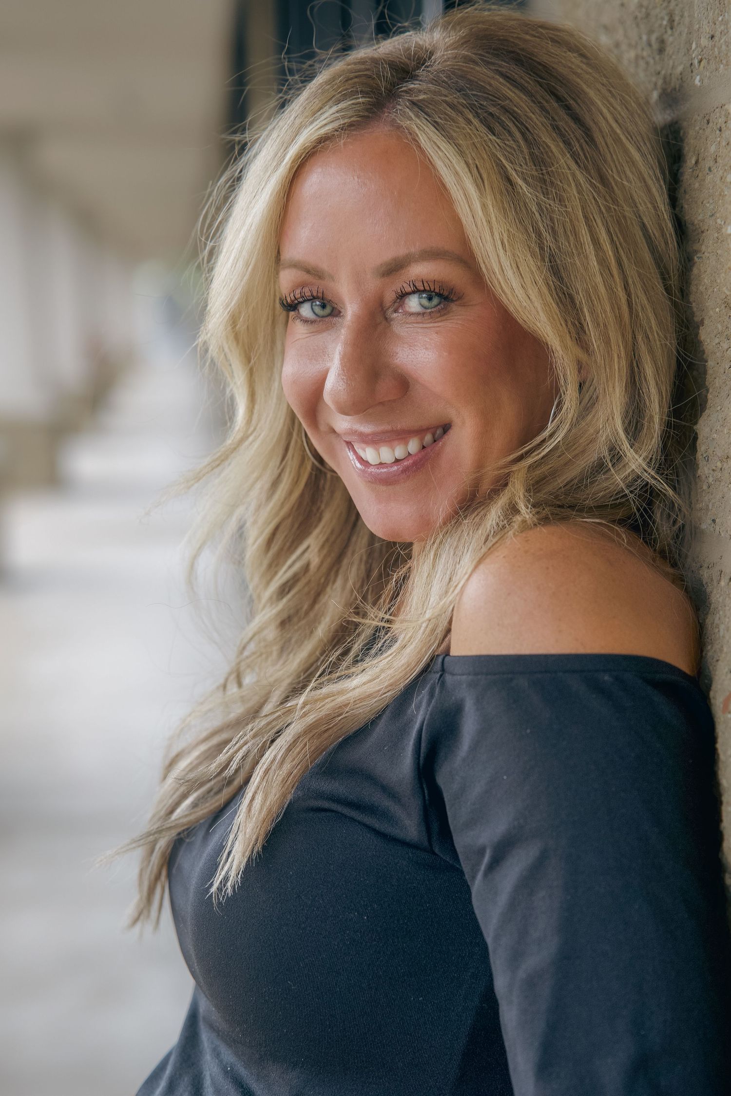 Smiling blonde woman with blue eyes leaning against a wall, wearing an off-shoulder dark top, with long wavy hair.