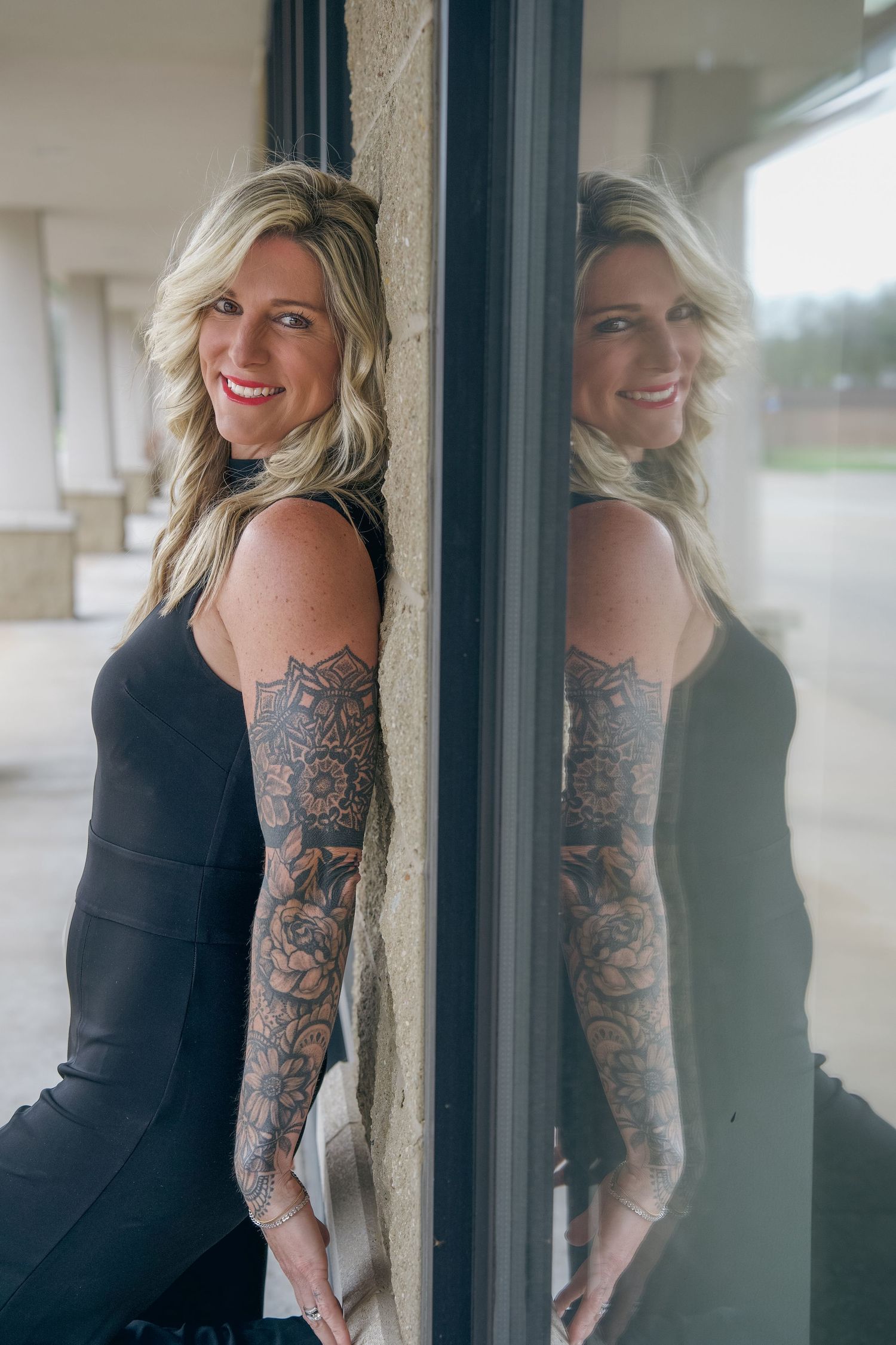 Blonde woman with full sleeve tattoos in black dress leaning against window with reflection visible.