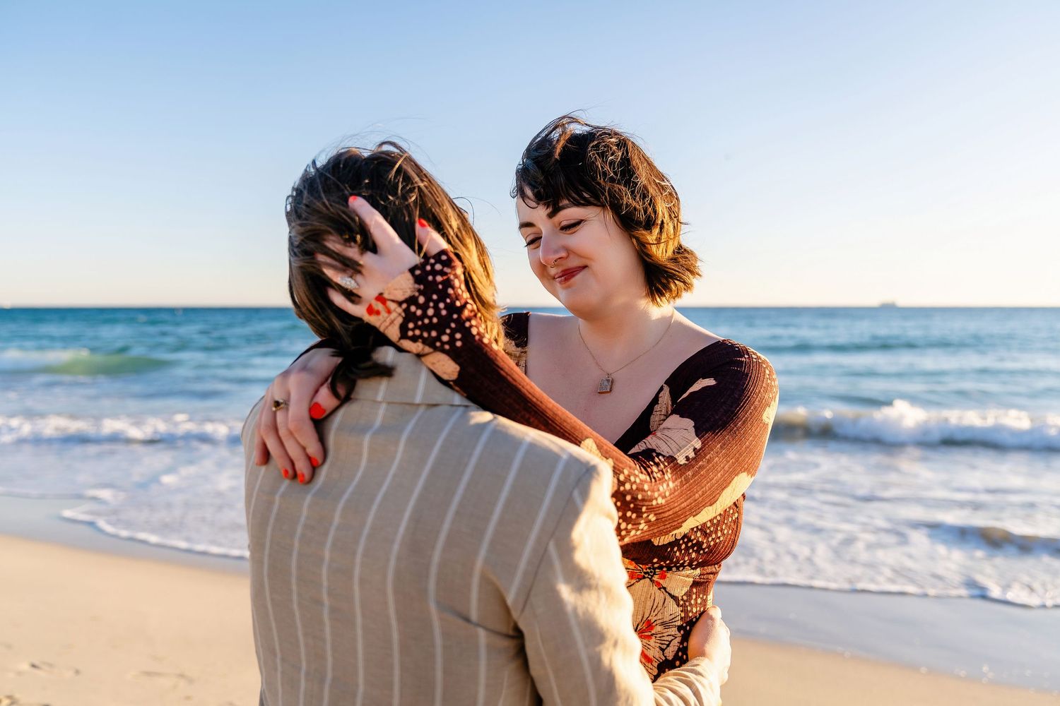 Couple embracing on a sunny beach with ocean waves, sharing a romantic moment at golden hour.