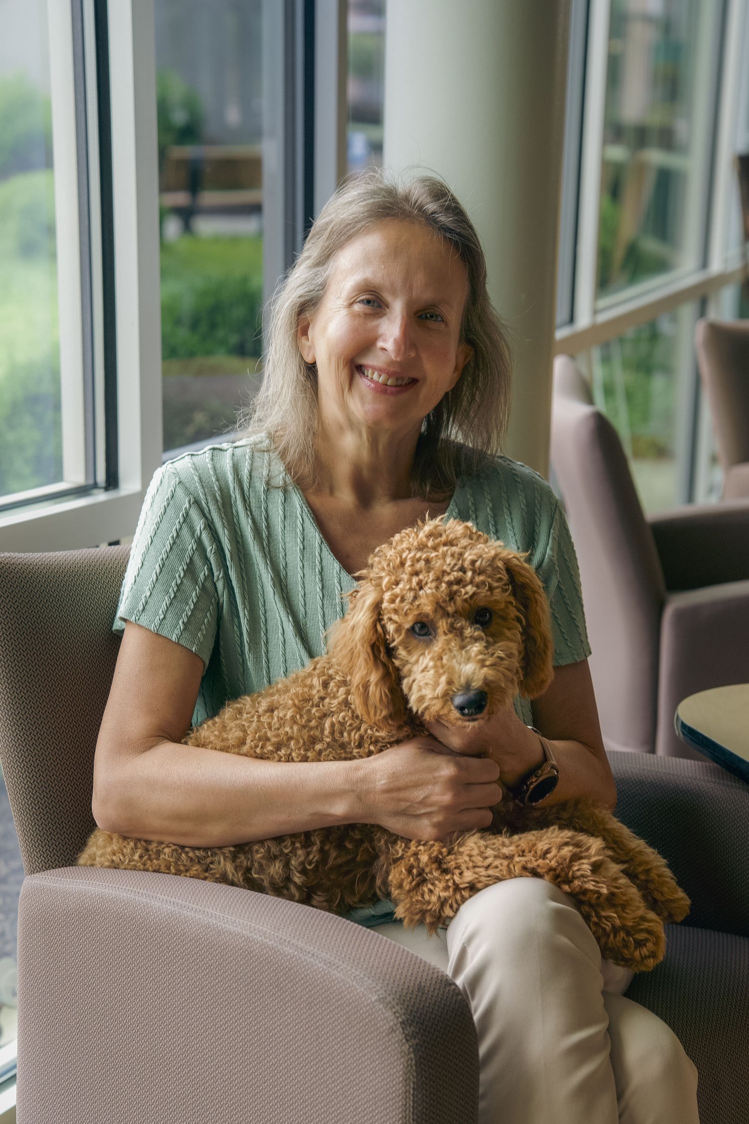Smiling older woman with gray hair holding a fluffy golden doodle dog while sitting in a chair near large windows.