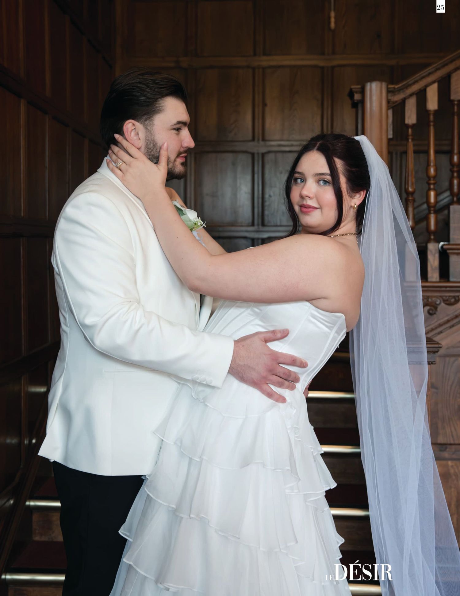 A bride in a white tiered gown and veil embraces a groom in a white tuxedo on elegant wooden staircase.