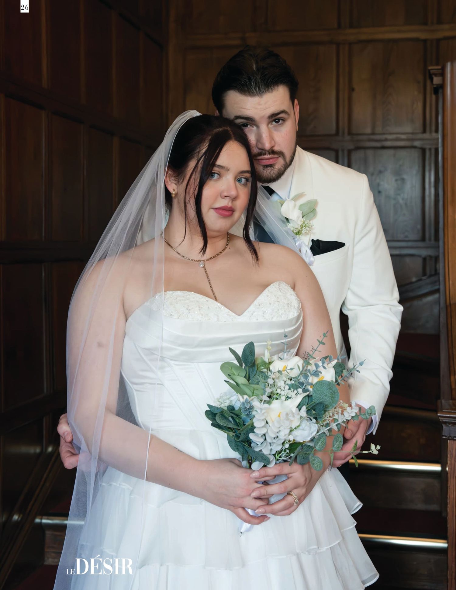 Bride in white strapless gown with veil holds floral bouquet as groom in white suit embraces her from behind.