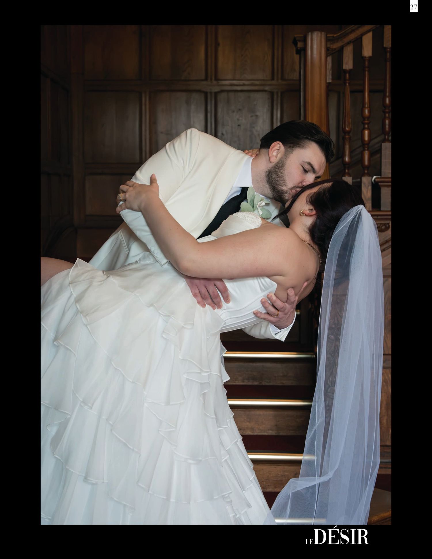 Groom dips and kisses bride in white gown and veil on elegant wooden staircase in romantic wedding portrait.