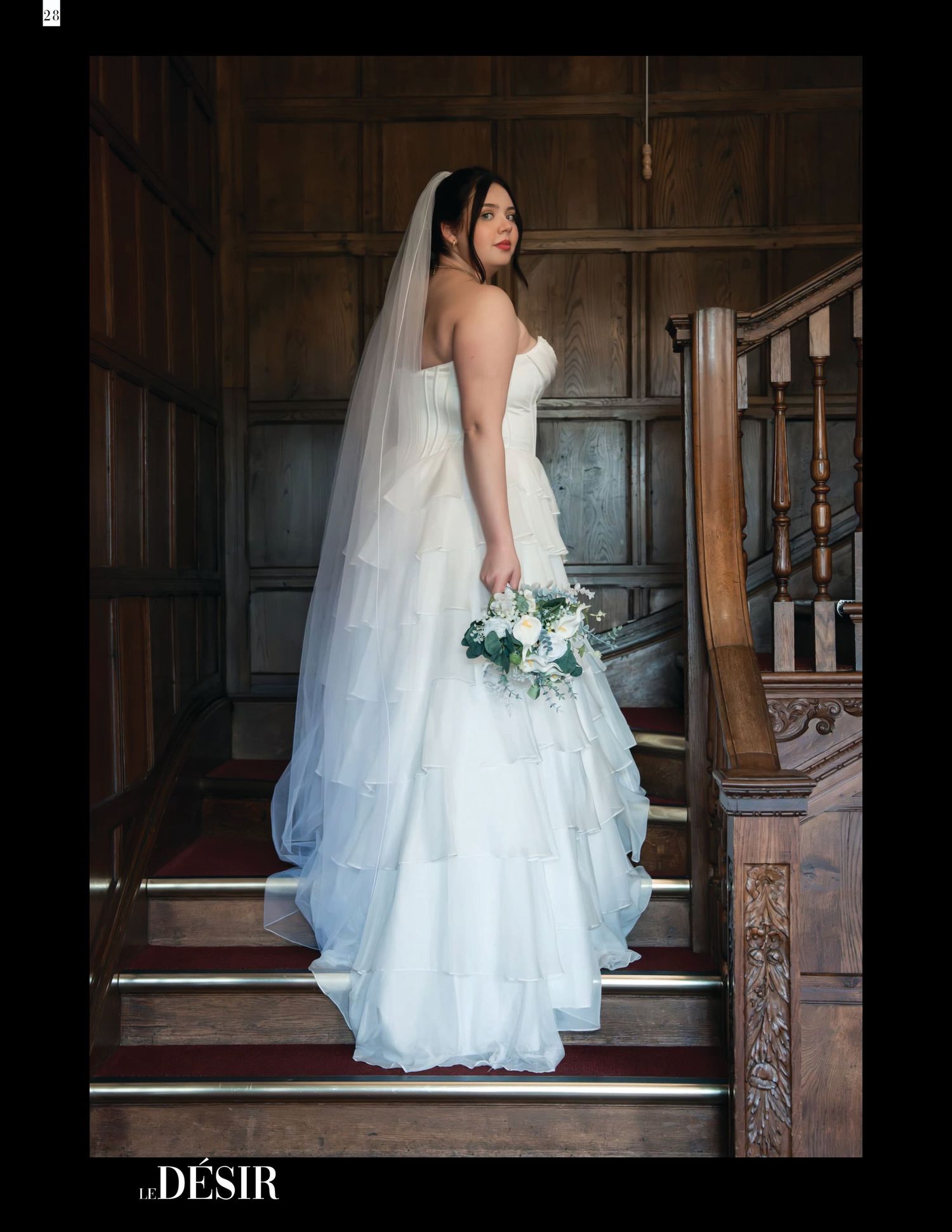 Bride in strapless white tiered gown with veil holds bouquet on wooden staircase in elegant venue.