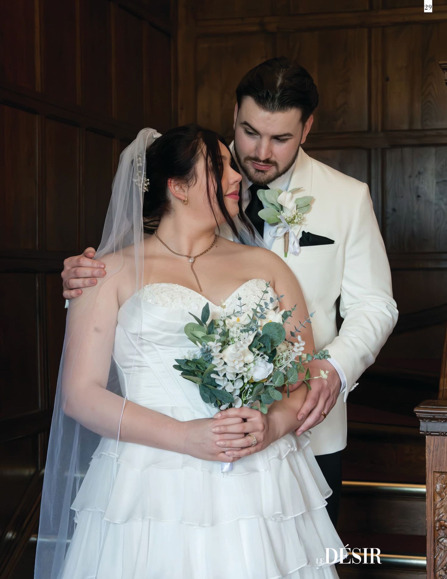 Elegant wedding couple embrace tenderly, bride holding white and green bouquet in white gown with veil.