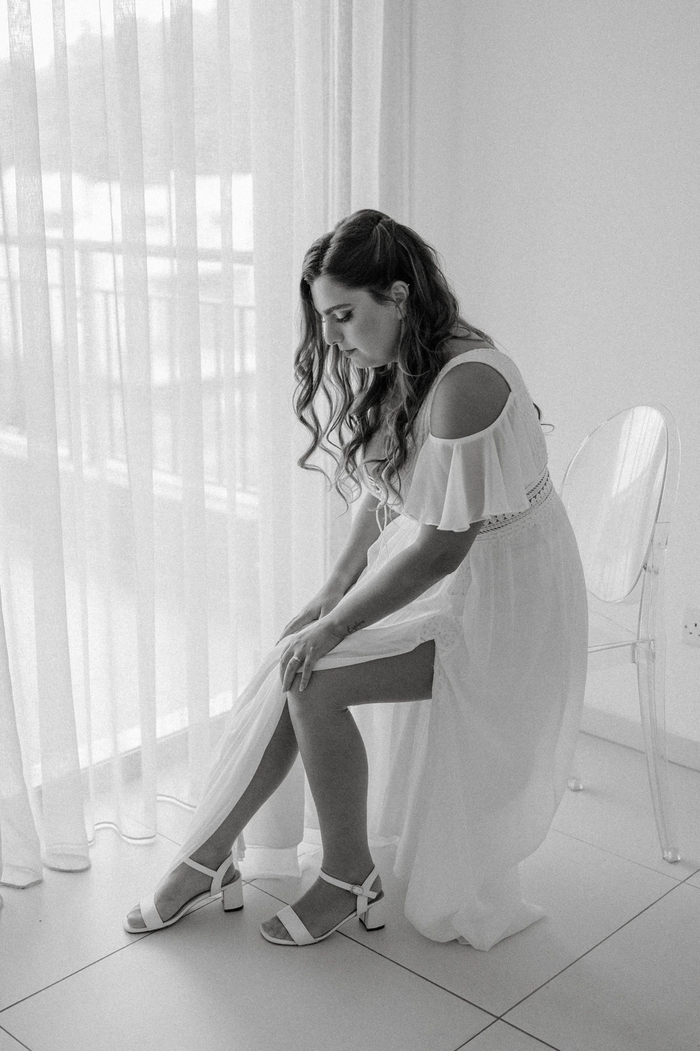 Bride in white off-shoulder dress adjusting heels while seated by sheer curtains, black and white wedding photo.