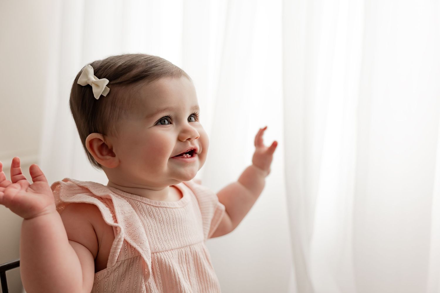 Smiling baby girl in pink ruffle romper with white bow, reaching up toward sheer white curtains.
