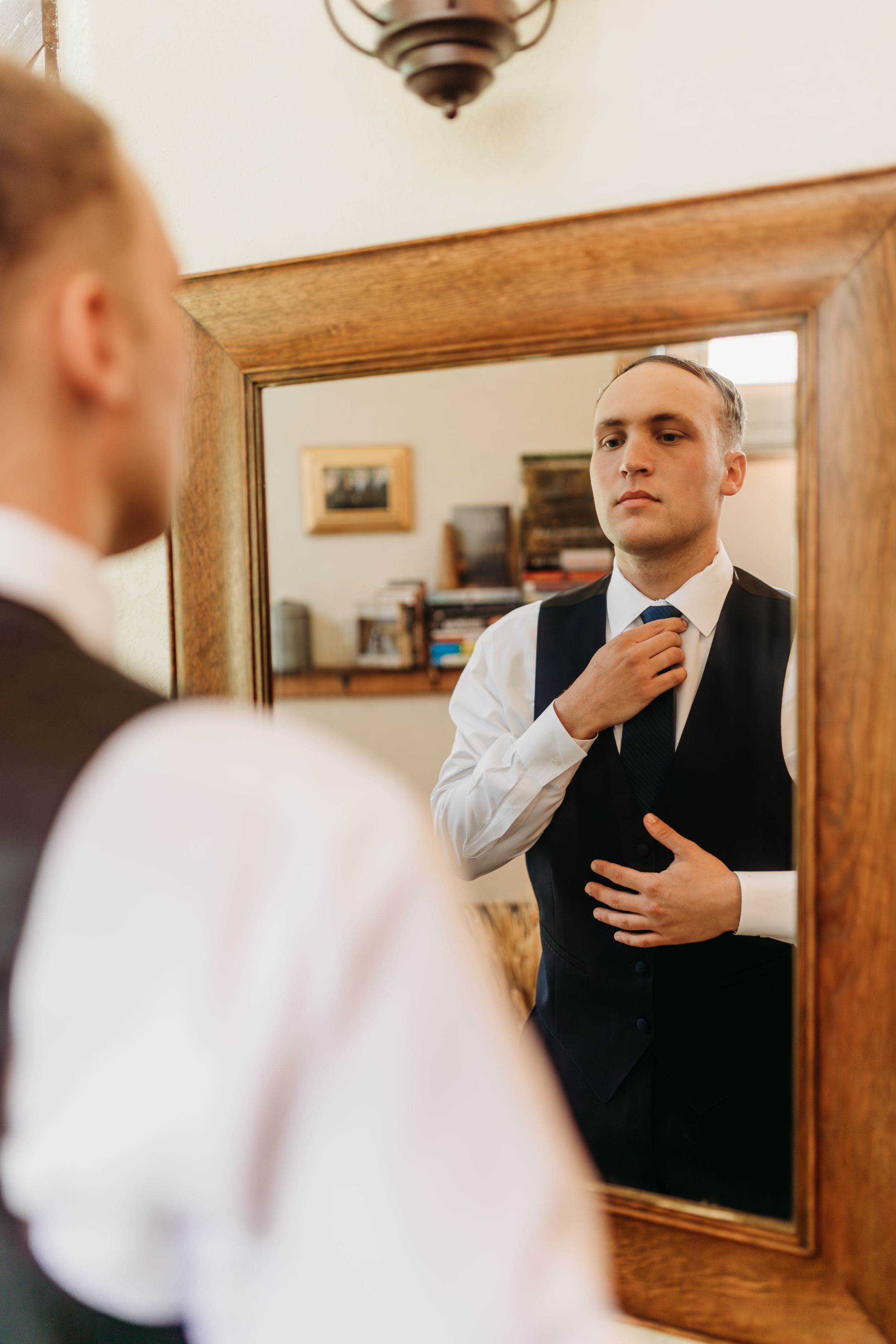 Groom adjusting his blue tie while looking at his reflection in a wooden framed mirror while getting ready.
