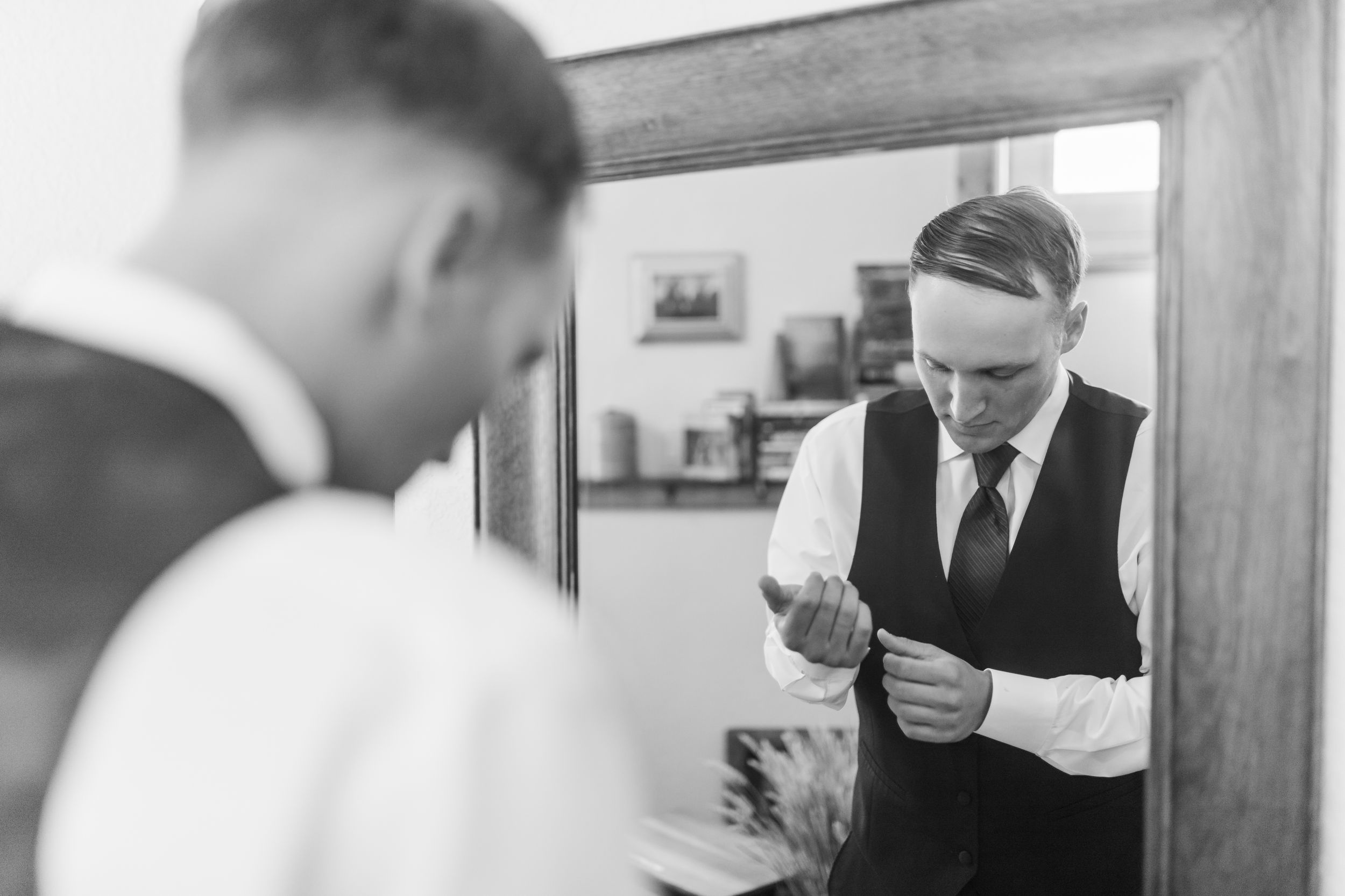 Black and white photo of a groom adjusting his cufflinks in front of a mirror while getting ready.