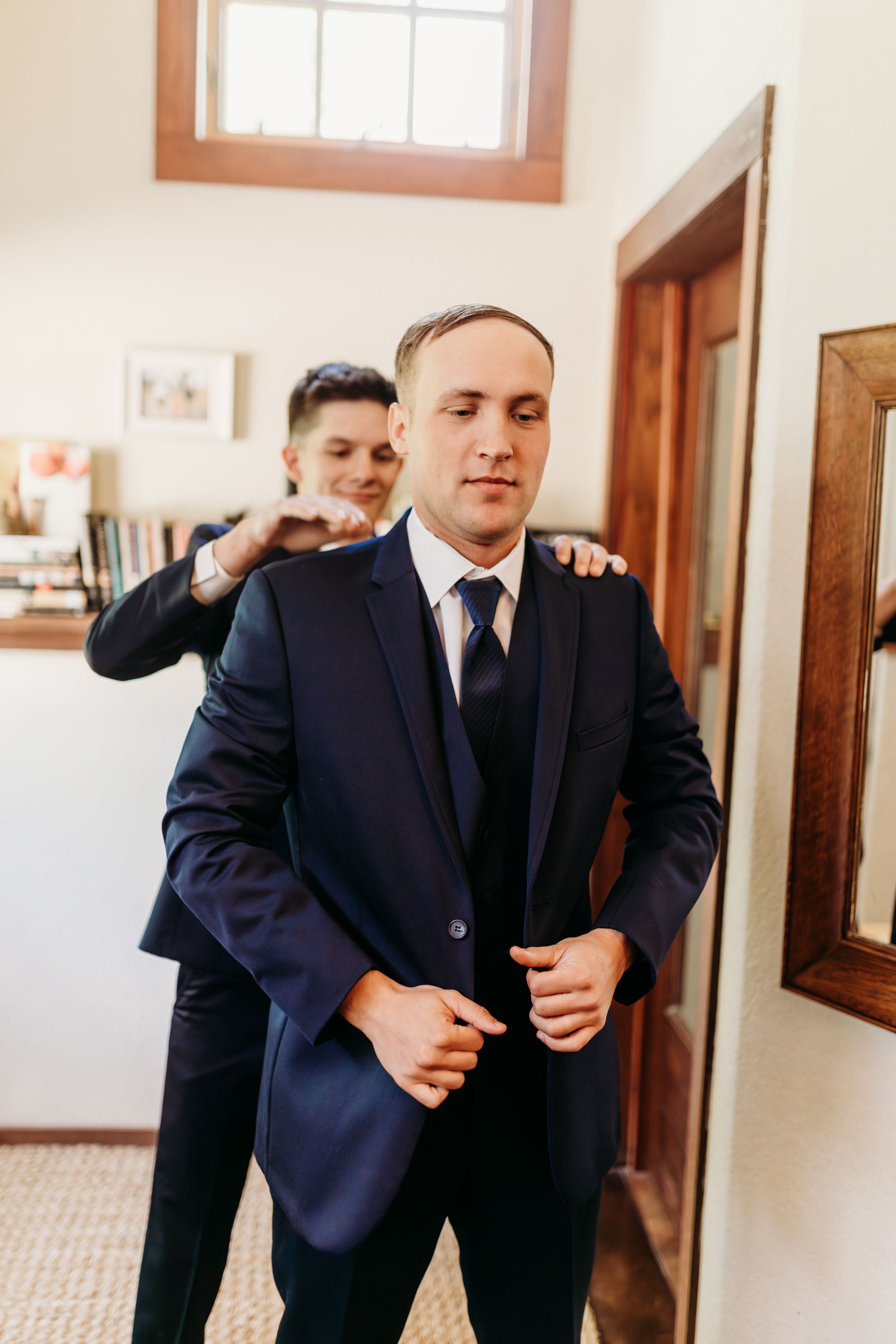 Groom buttoning his navy suit jacket with groomsman adjusting his collar before wedding ceremony.