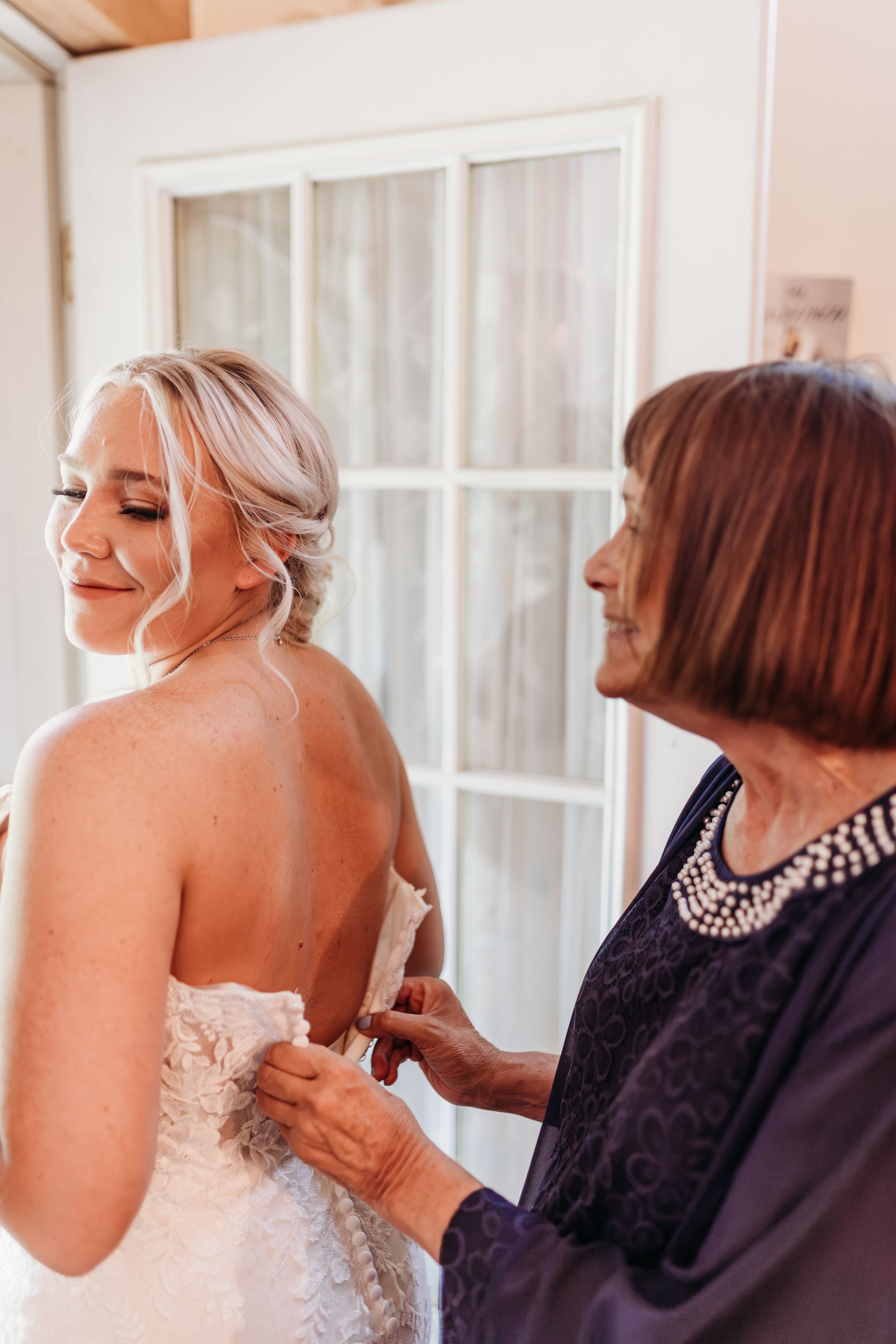 Older woman helping smiling blonde bride button the back of her lace wedding dress.