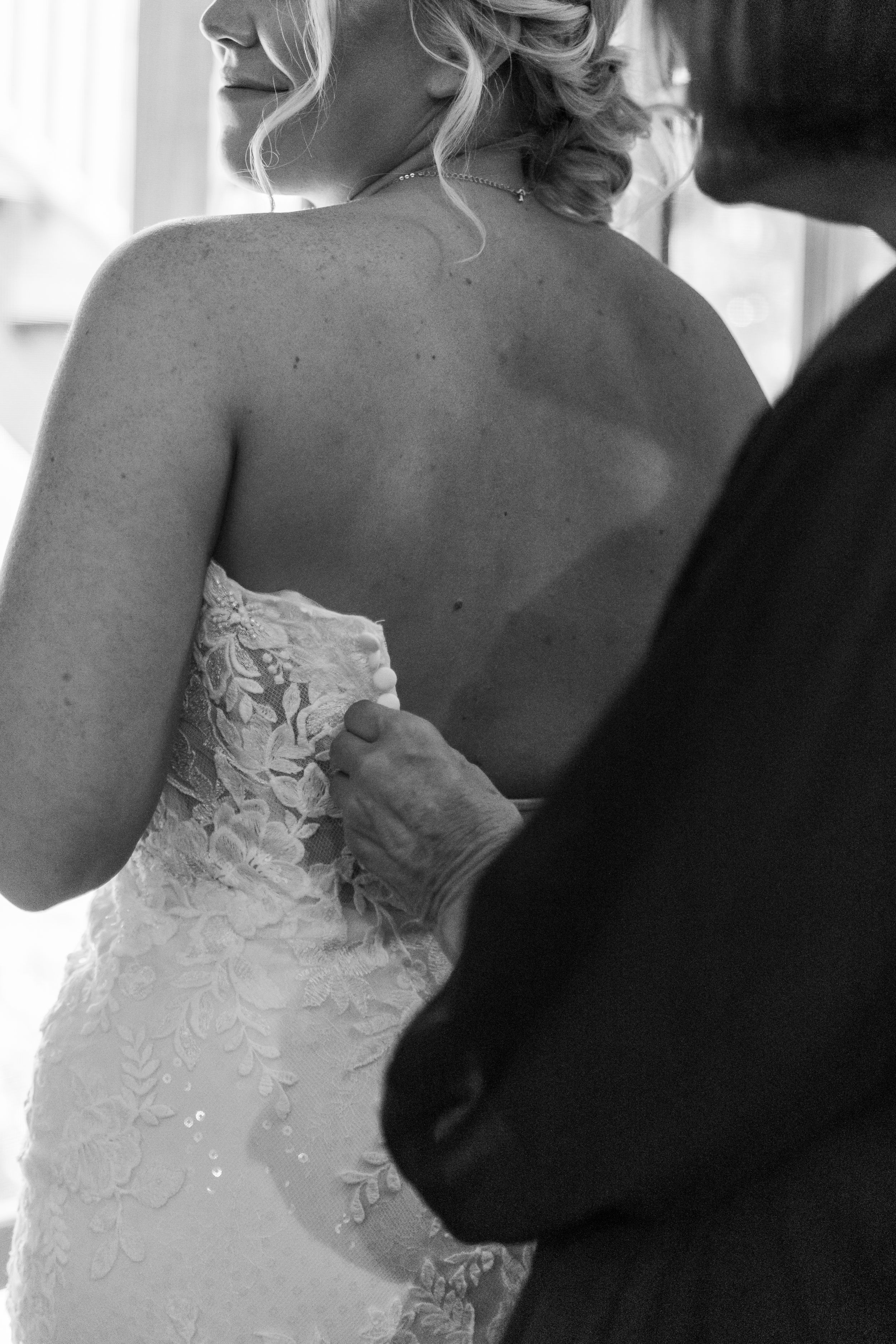 Black and white photo of someone buttoning the back of a brides lace wedding dress.