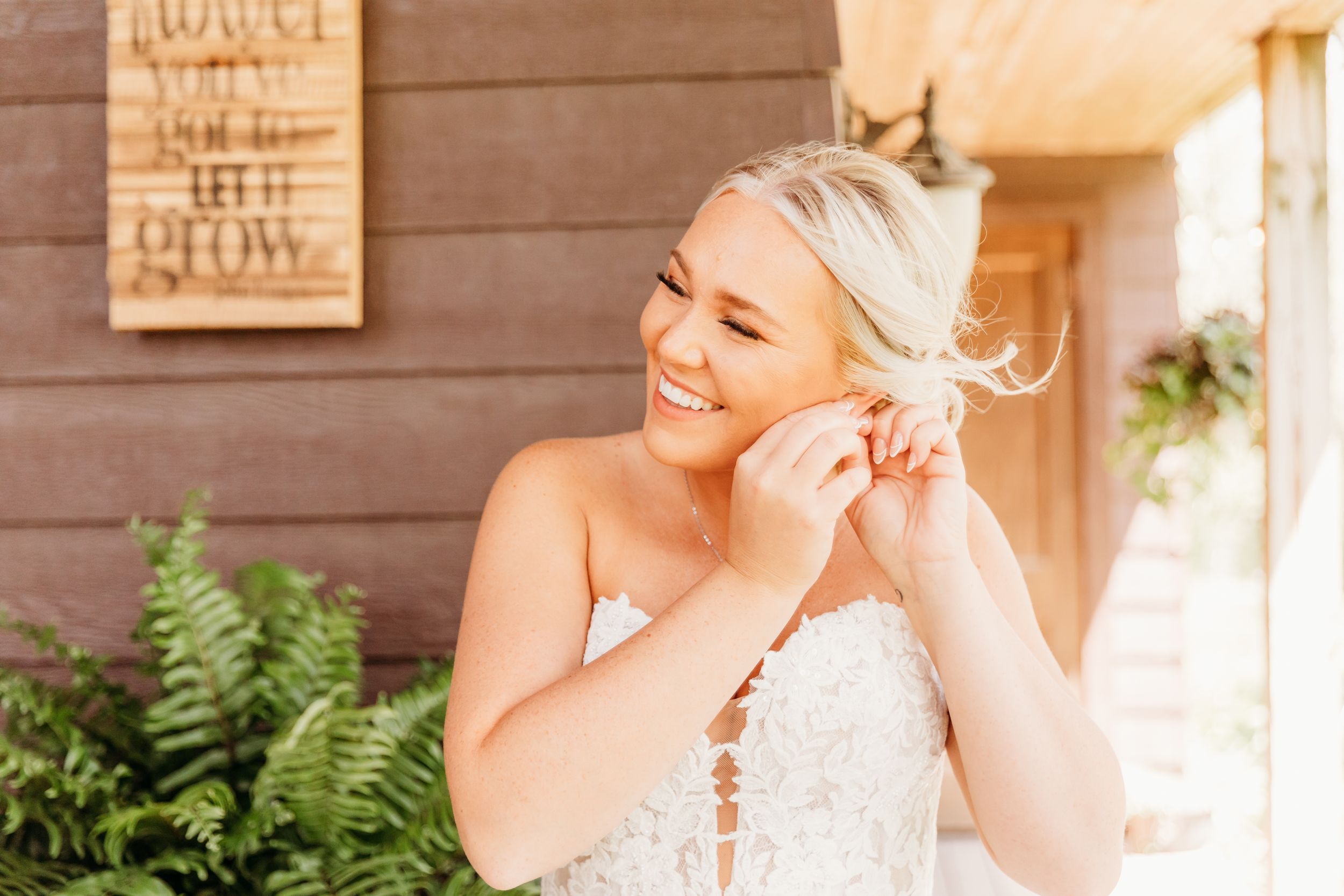 Smiling blonde bride in lace wedding dress adjusting earring outdoors near rustic wood building with ferns.