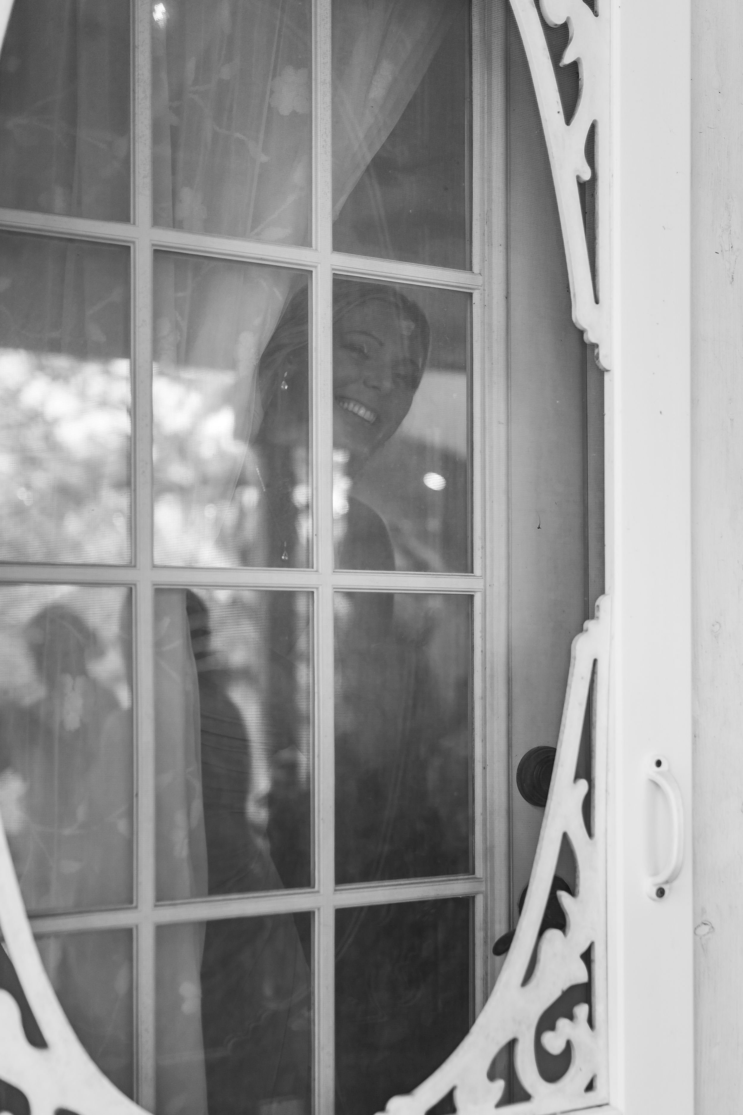 Black and white photo of a smiling person seen through an ornate white French door with glass panes.