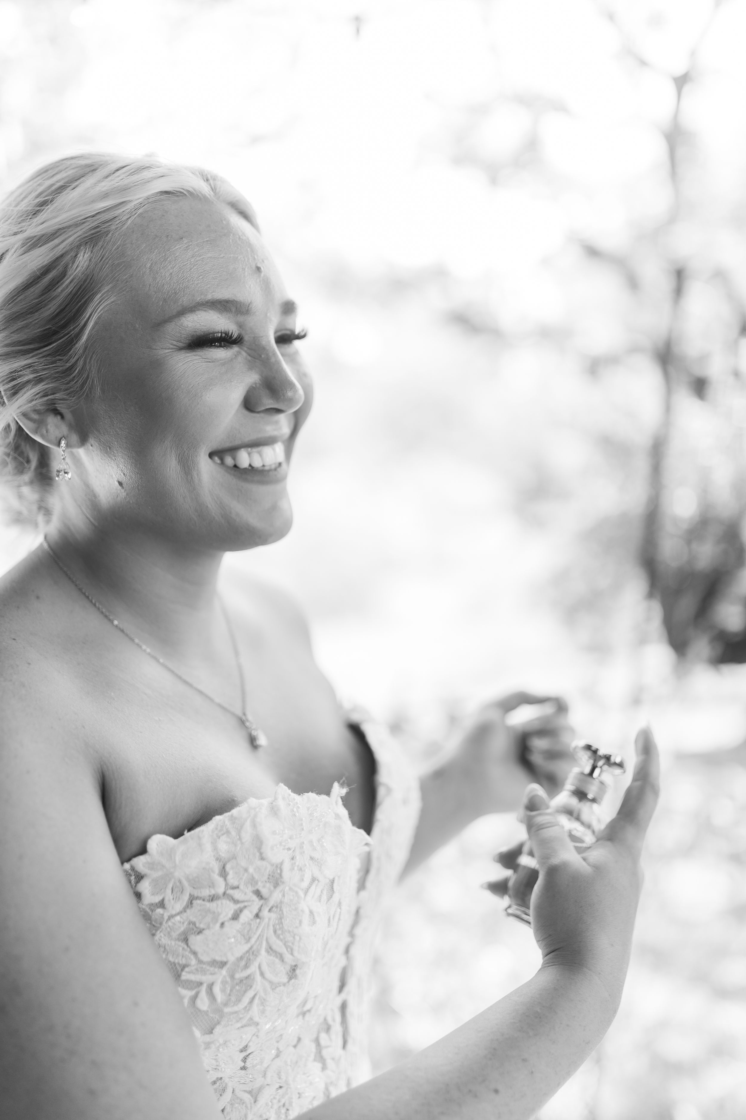 Smiling bride in lace strapless gown applying perfume outdoors in black and white wedding photo.
