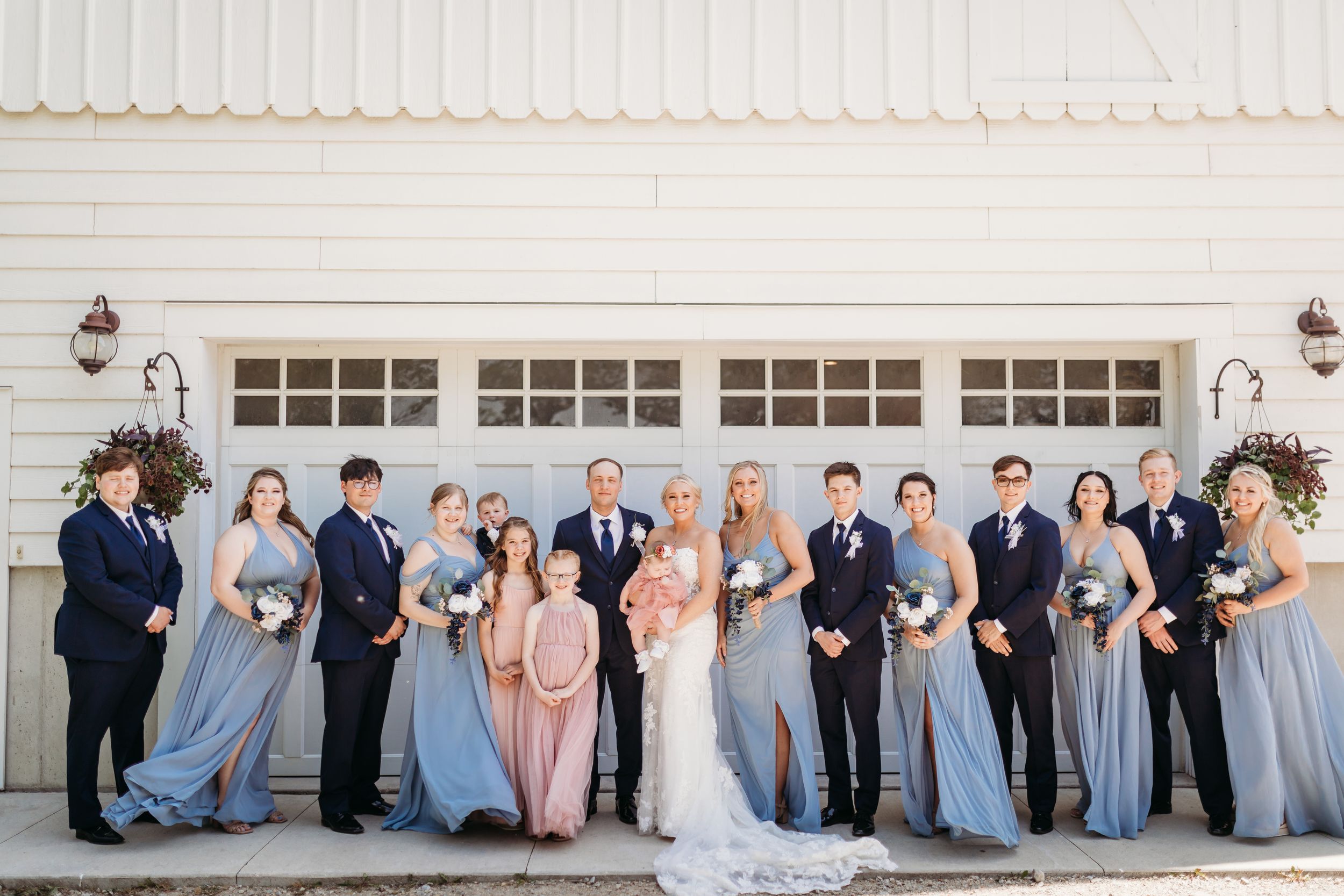 Wedding party poses outside white barn, bridesmaids in dusty blue gowns flanking bride and groomsmen in navy suits.