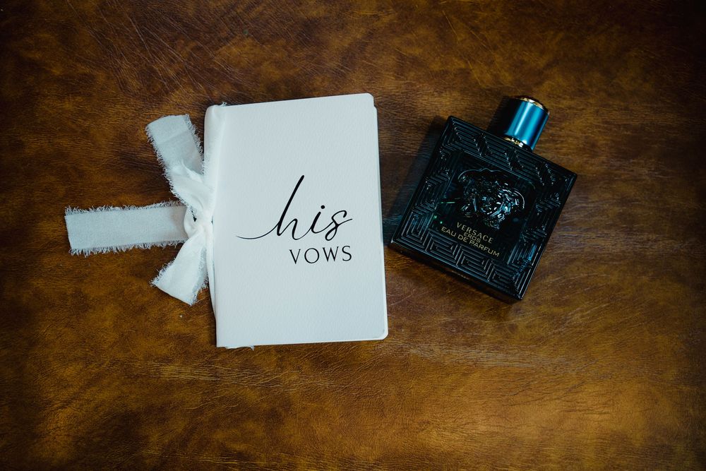 His vows book with white ribbon bow beside Versace Eros cologne bottle on rustic brown leather surface.