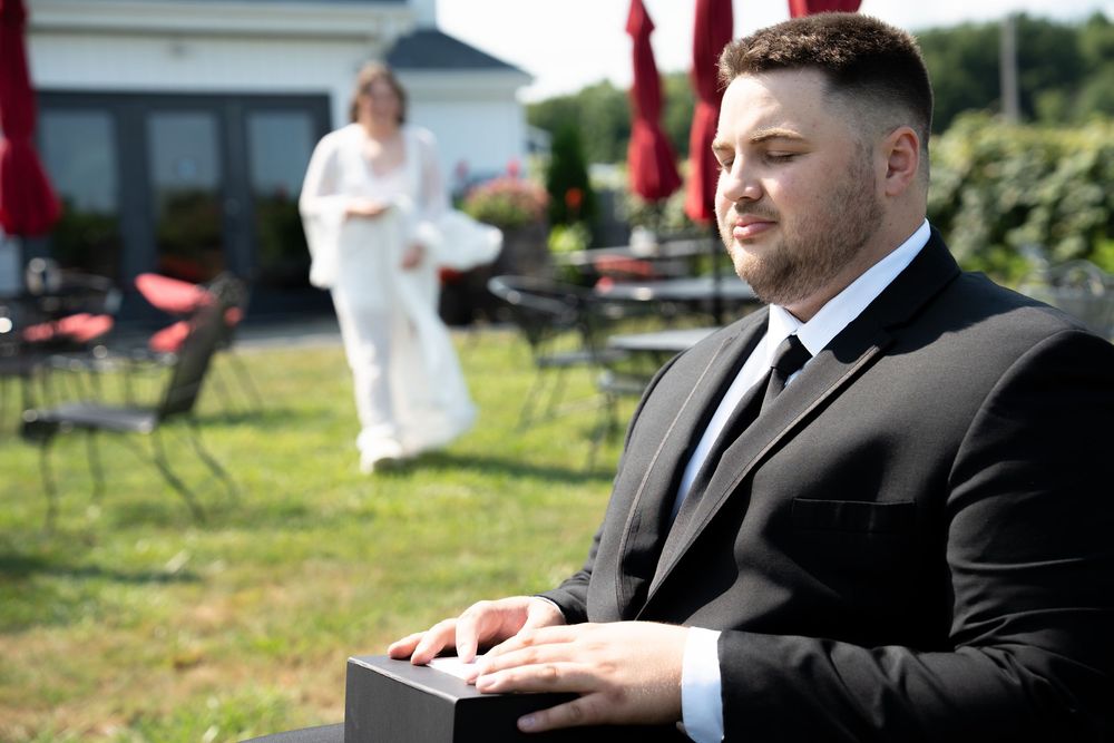 Groom in black suit holding box outdoors at wedding venue with guests in background.