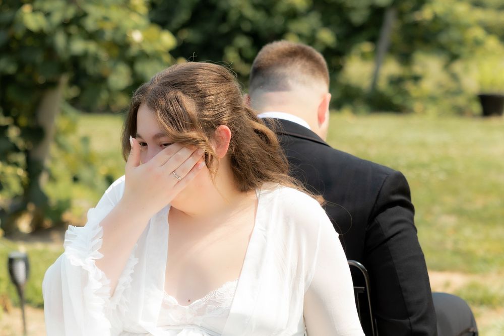 Emotional bride covering her mouth during outdoor wedding ceremony seated beside groom in black suit.