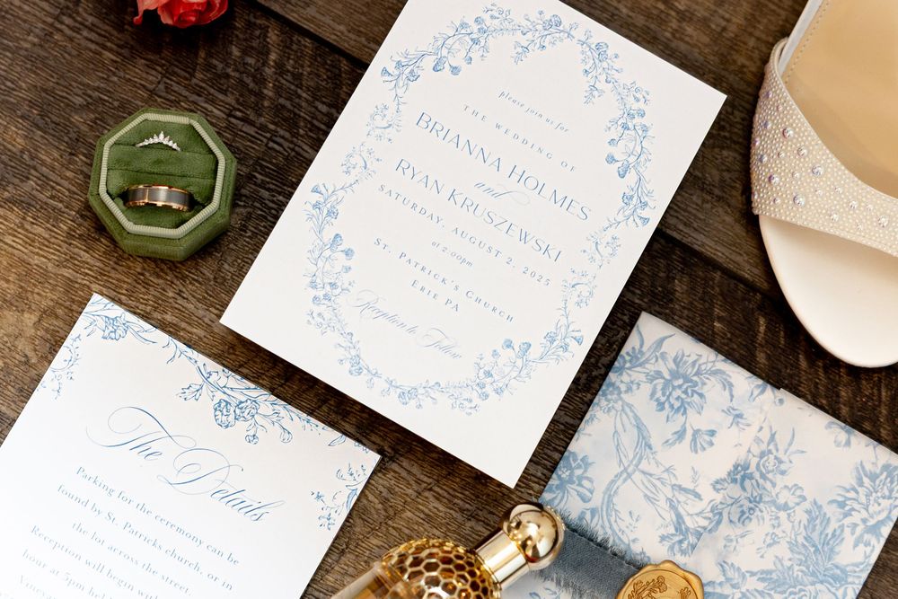Elegant blue and white floral wedding invitation suite with rings and details card on rustic wood.