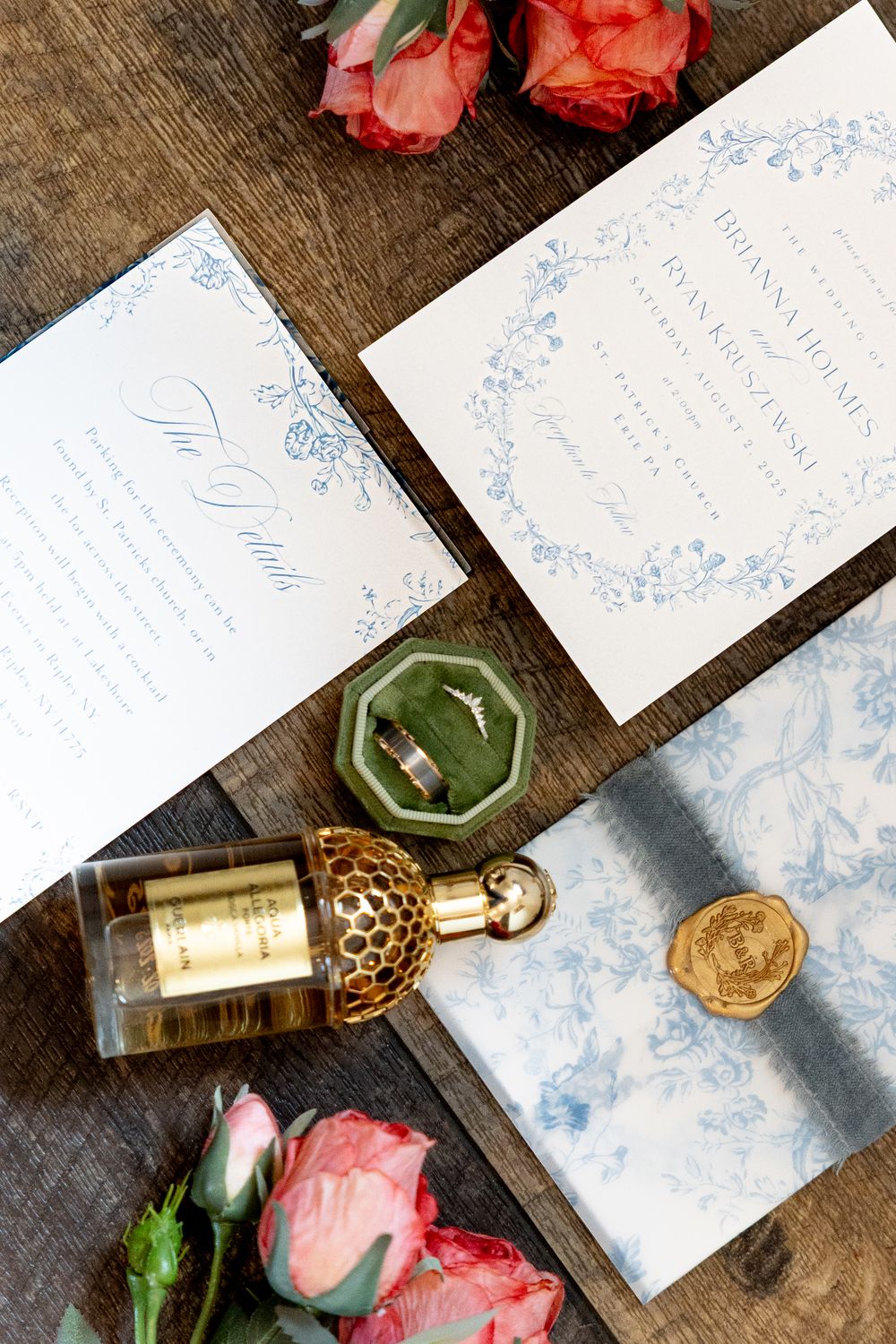 Elegant wedding flat lay with blue floral invitations, diamond ring in velvet box, perfume bottle, and pink roses on wood.