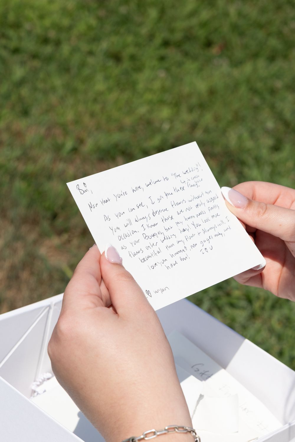Hands holding a handwritten note card outdoors with green grass in the background near a white gift box.