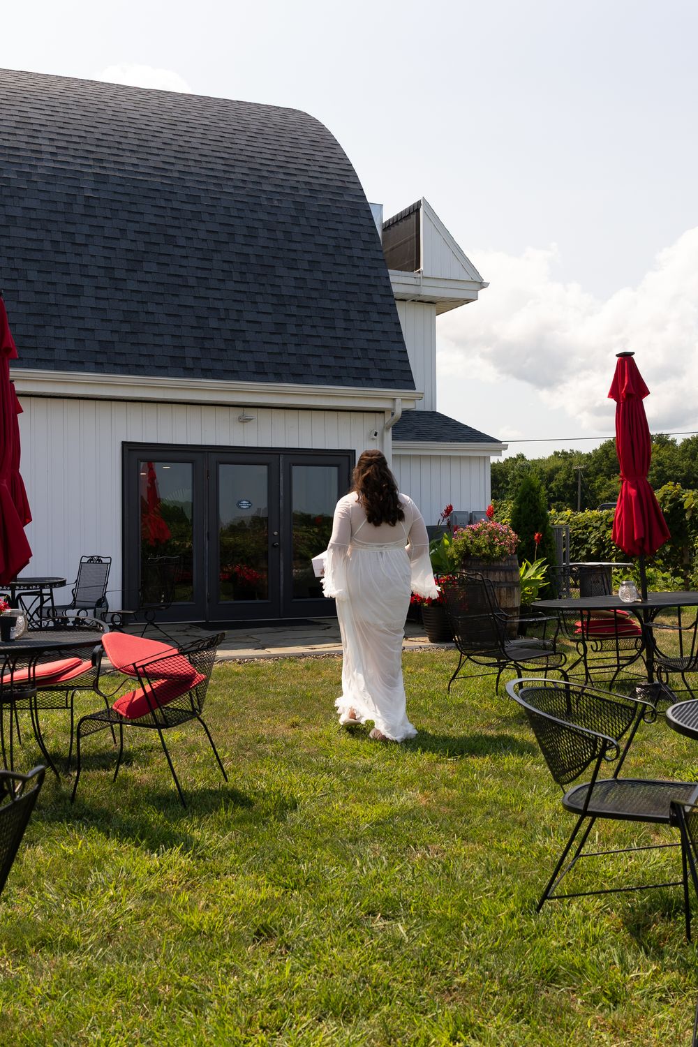 Bride in white gown walks toward white barn venue with red patio umbrellas and black metal chairs on green lawn.